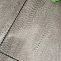 Clean House Jaworski Przemysław - Jasnoszara podłoga z paneli imitujących drewno, ułożonych ukośnie, z fragmentem zielonego koszyka w prawym górnym rogu. Widoczne fugi między panelami.