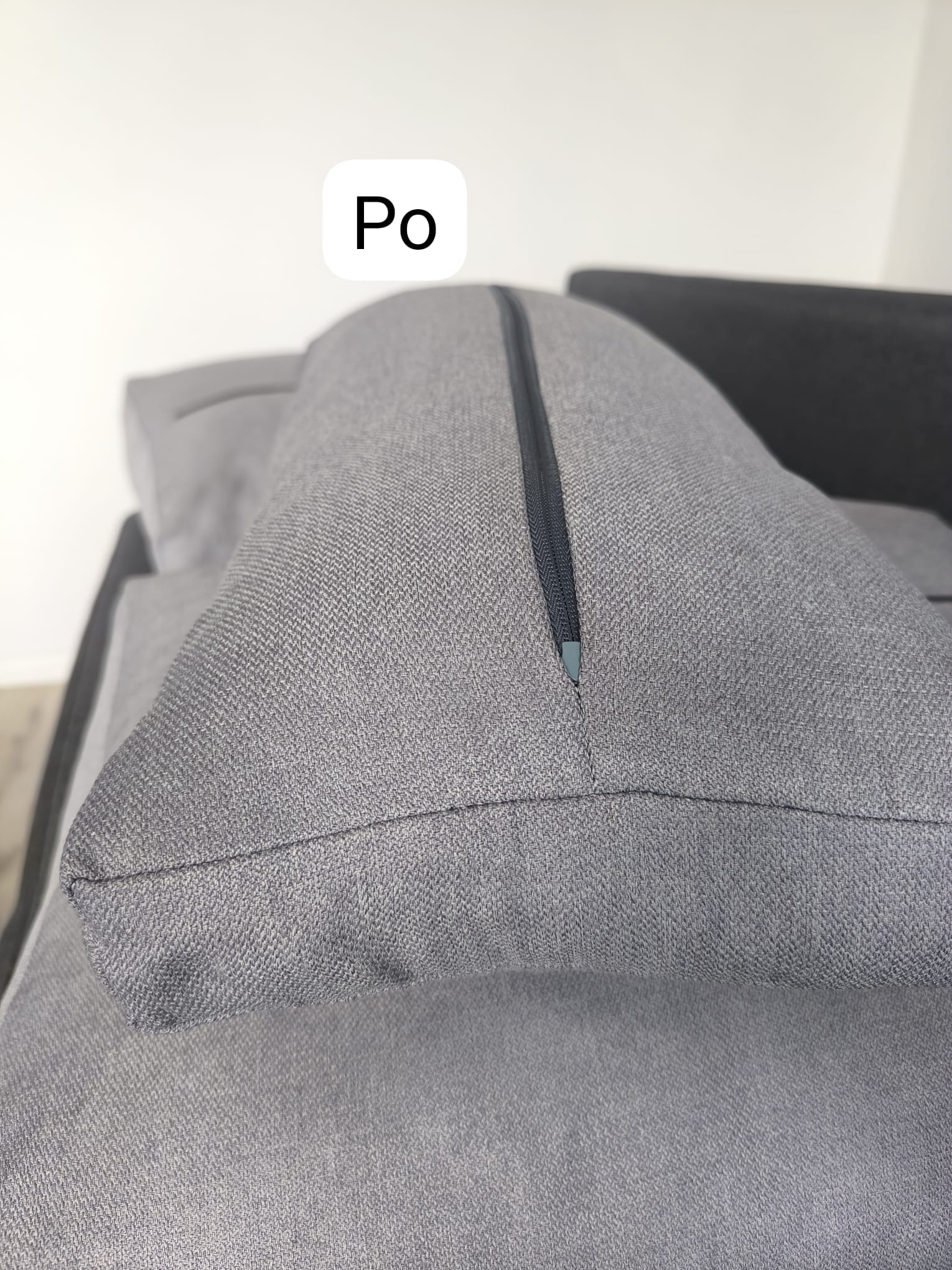 Szara poduszka z zamkiem błyskawicznym, leżąca na szarej kanapie o wyraźnej teksturze materiału.