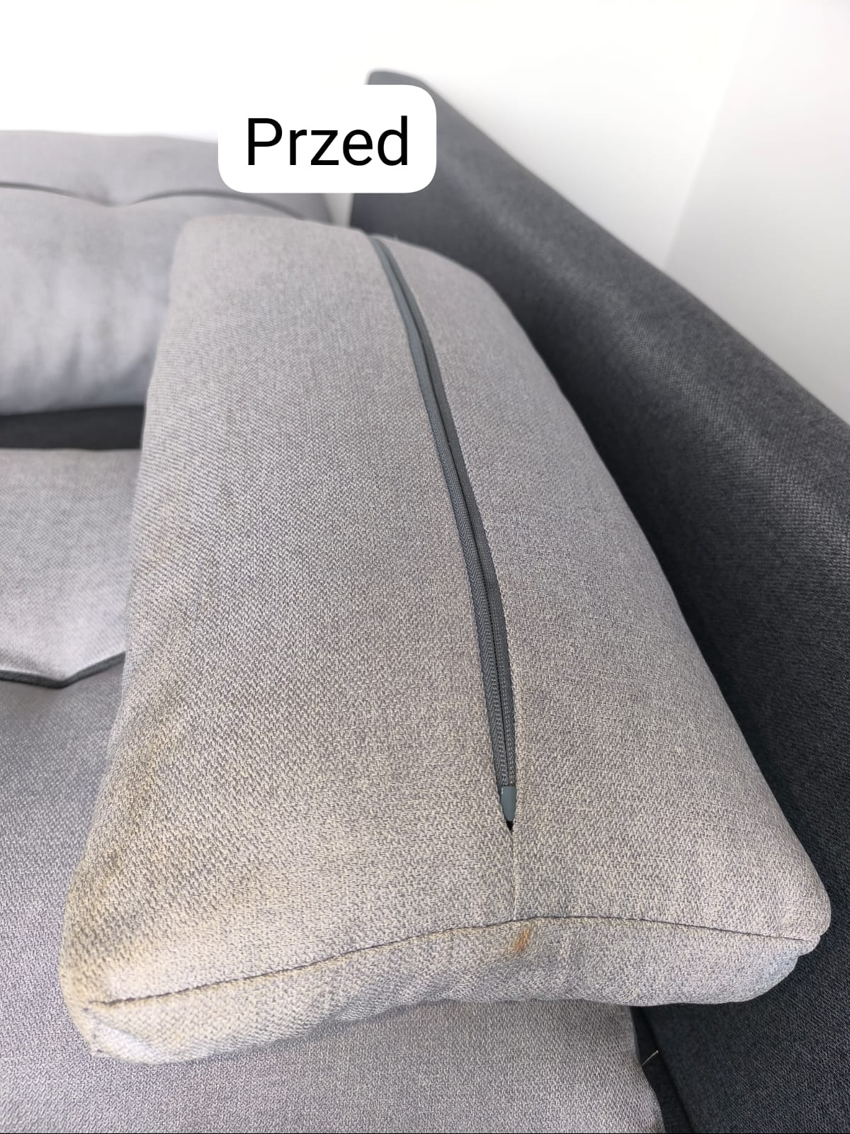 Szara poduszka kanapy z widocznymi plamami i zamkiem błyskawicznym przed praniem.