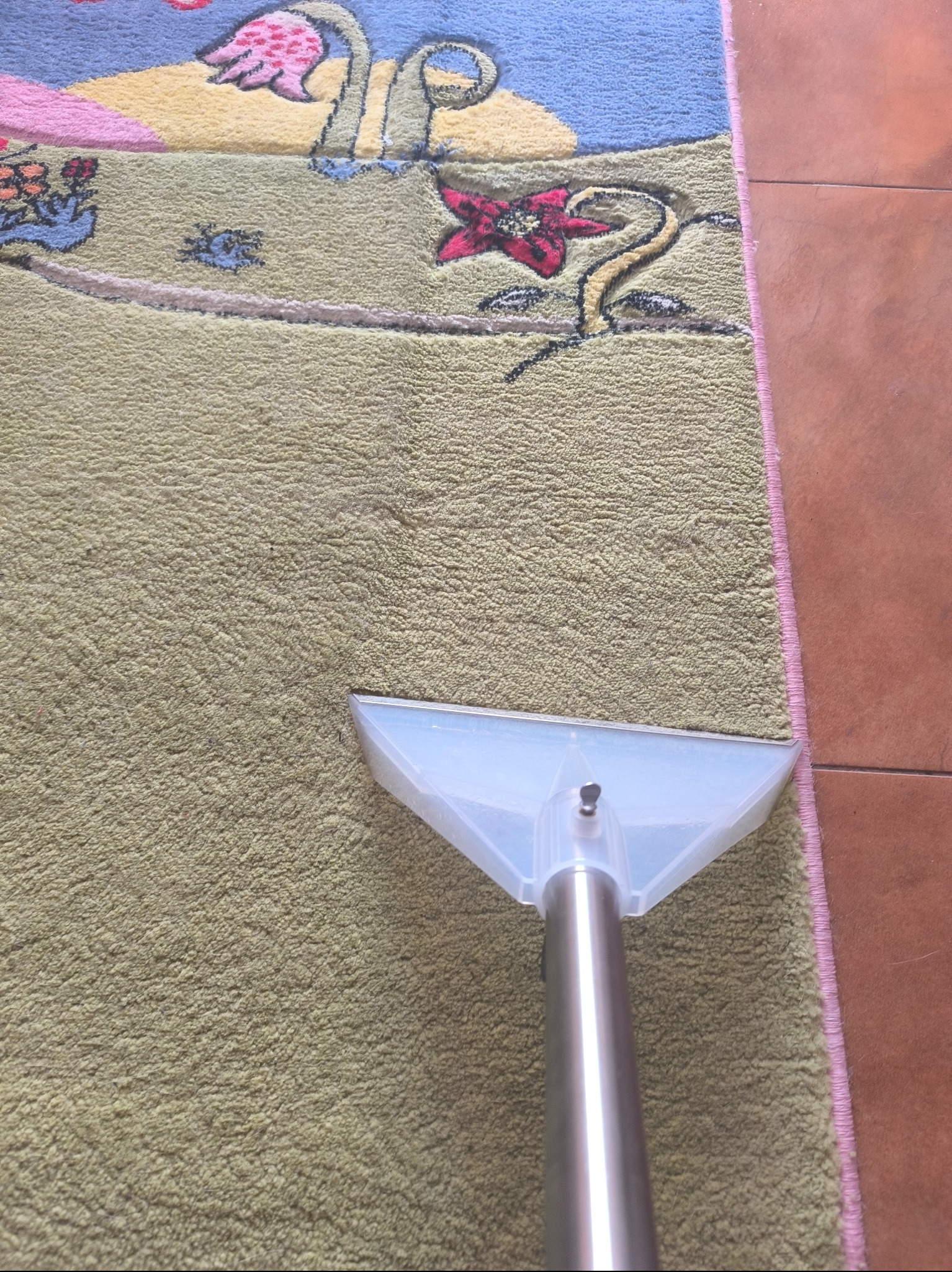 Czyszczenie dywanu z dziecięcym wzorem za pomocą odkurzacza piorącego z przezroczystą końcówką i metalową rurą, widoczny fragment brązowej podłogi.