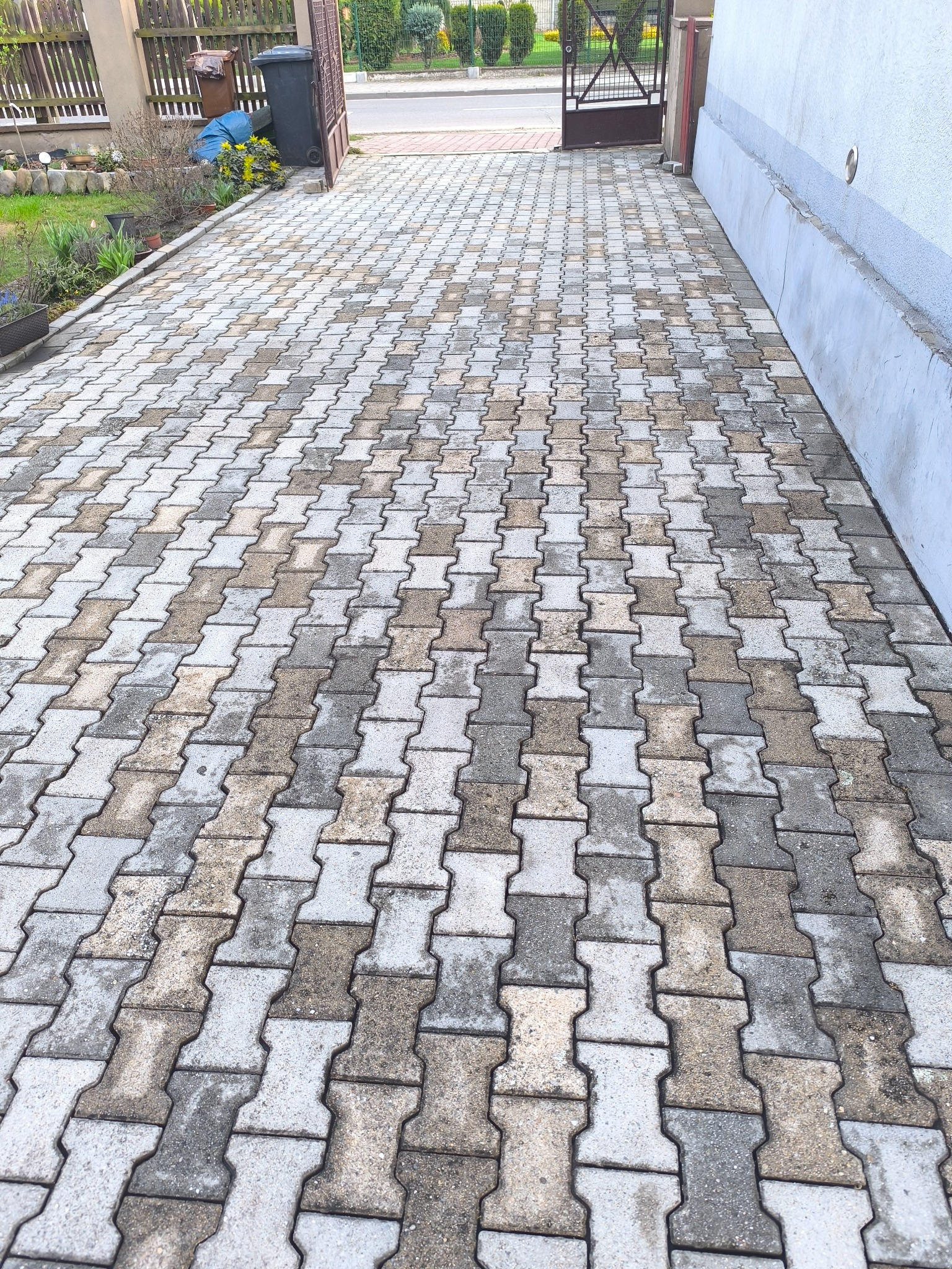 Ułożona kostka brukowa w kształcie litery I, prowadząca do bramki i chodnika. Część kostki jest jeszcze zabrudzona, co sugeruje potrzebę czyszczenia.