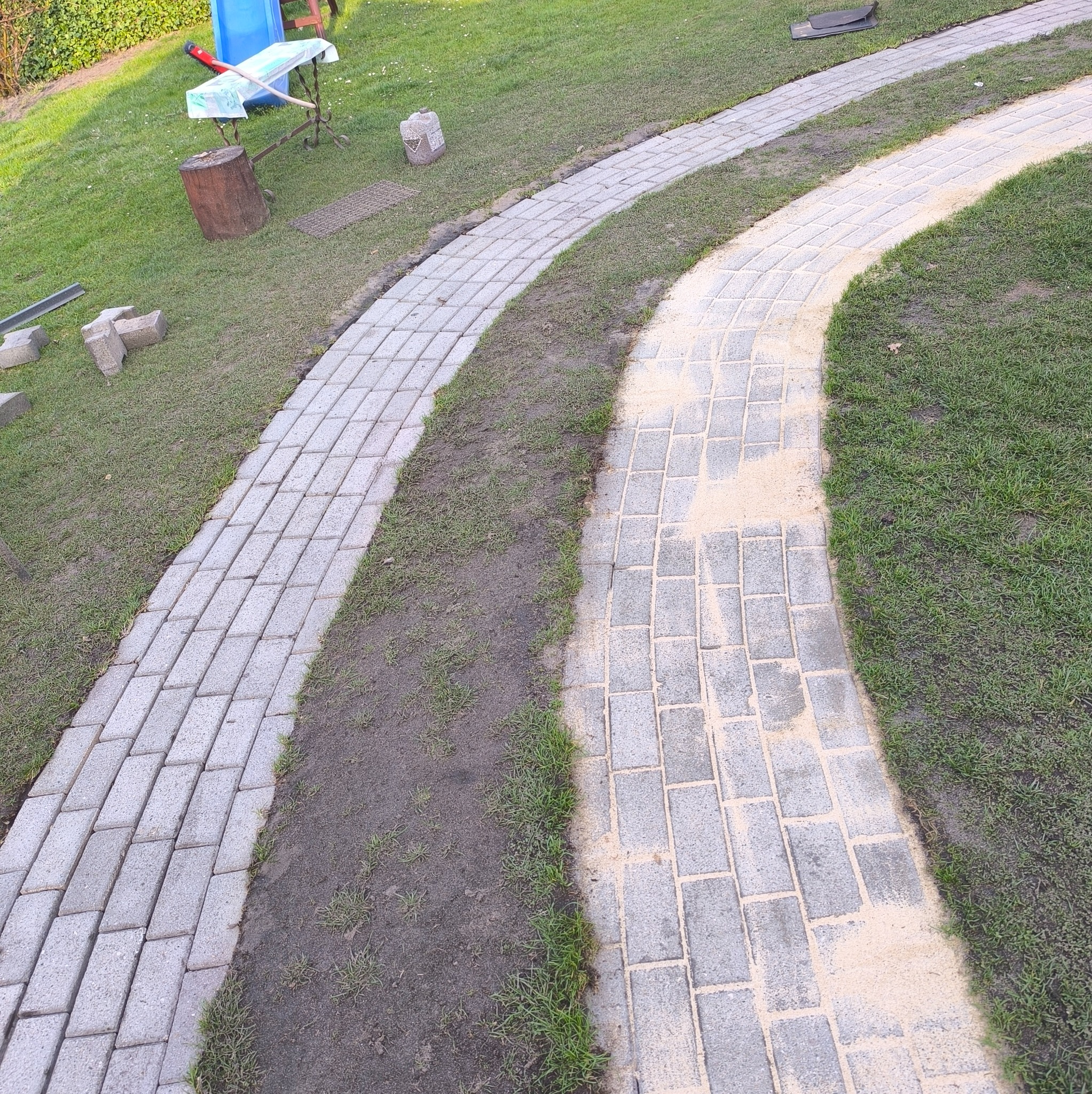 Ścieżka z kostki brukowej wijąca się przez trawnik. Część ścieżki jest już ułożona, a część przygotowana pod układanie. Widoczne narzędzia i materiały budowlane w tle.