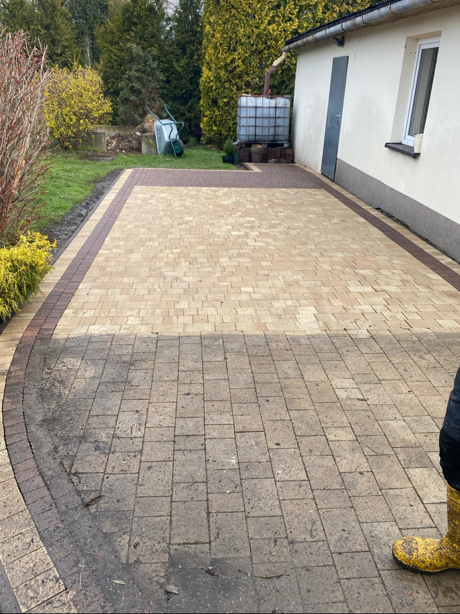 Ułożona kostka brukowa na podjeździe, z widocznym fragmentem czyszczonej powierzchni. W tle budynek z białymi ścianami i zielona roślinność.
