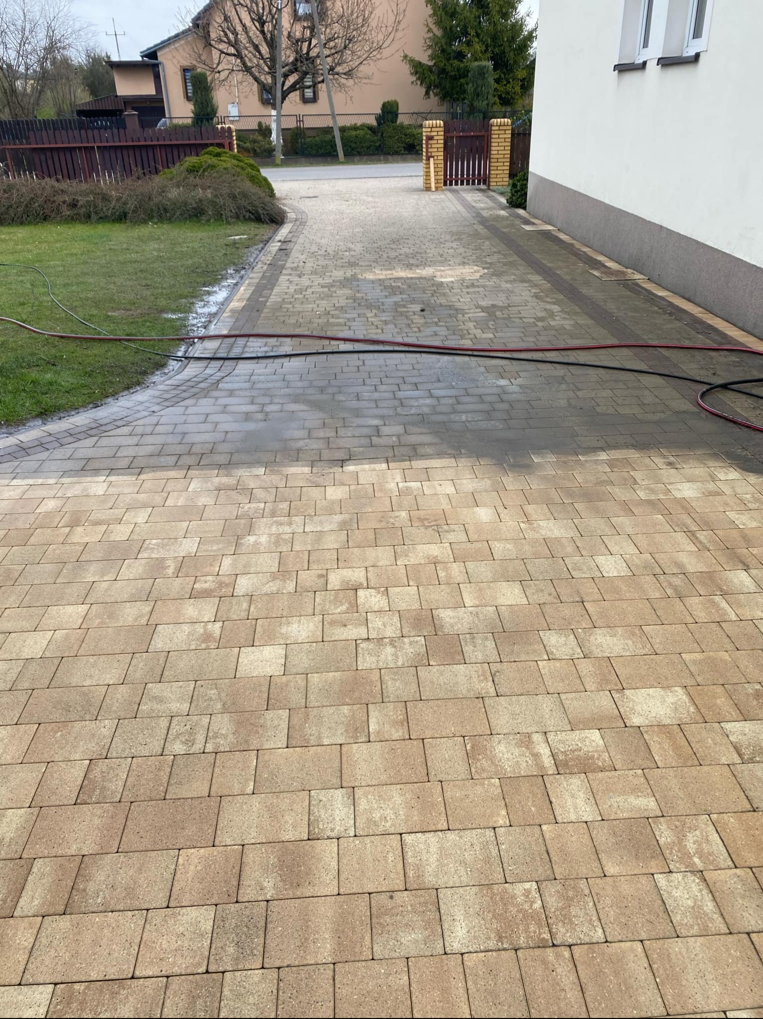 Częściowo umyta kostka brukowa na podjeździe, widoczna różnica w kolorze. W tle trawnik, budynek i brama wjazdowa. Wąż ciśnieniowy na kostce.