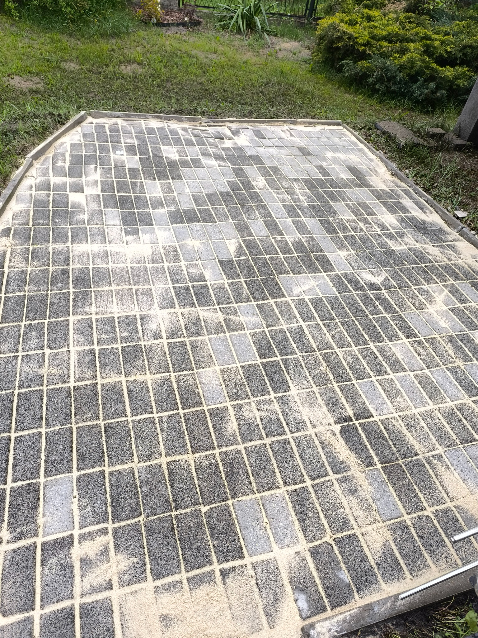 Ułożona kostka brukowa w odcieniach szarości, wypełniona piaskiem, tworząca równą powierzchnię na zewnątrz. Widoczne obrzeża i trawa w tle.