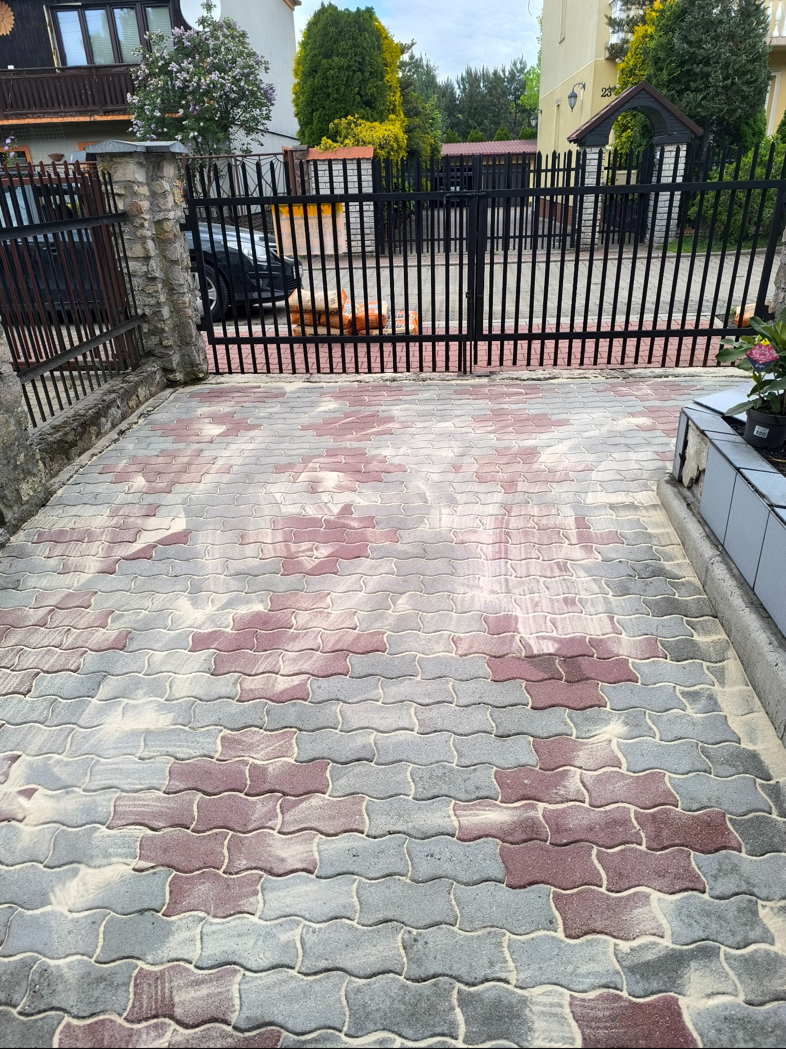 Wzorzysta kostka brukowa w odcieniach szarości i czerwieni, częściowo pokryta piaskiem, z widocznym fragmentem ogrodzenia i zieleni w tle.