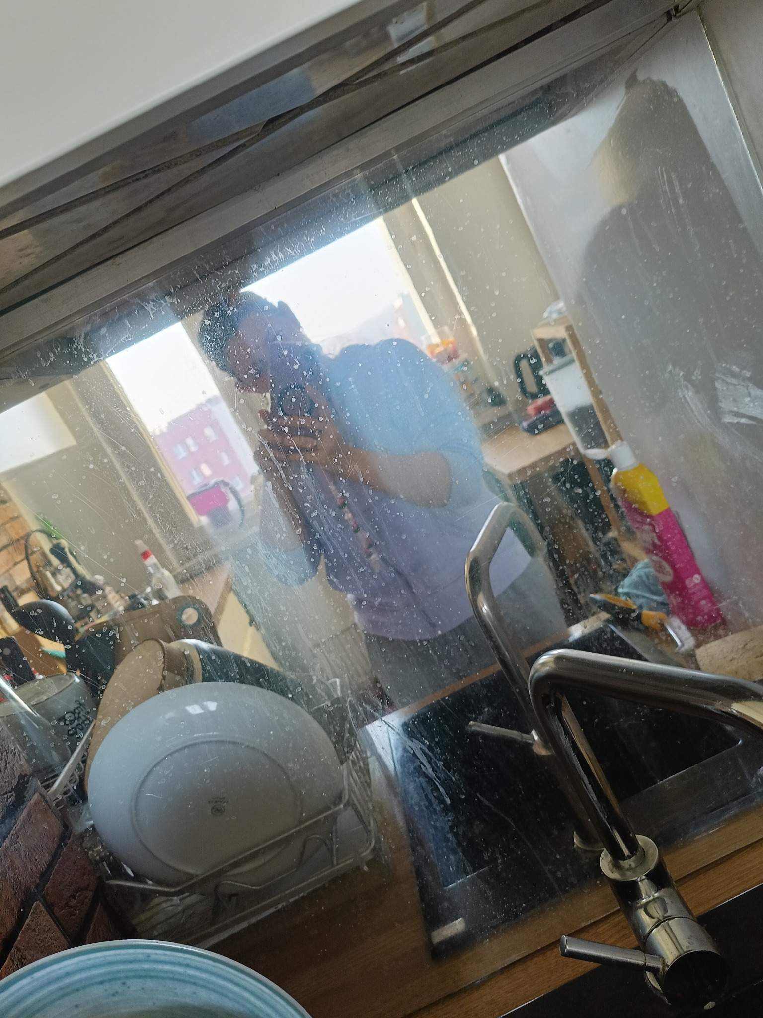 Zlewozmywak z naczyniami widziany przez brudne okno. Widoczne zacieki i plamy na szybie. W tle odbicie osoby robiącej zdjęcie telefonem.
