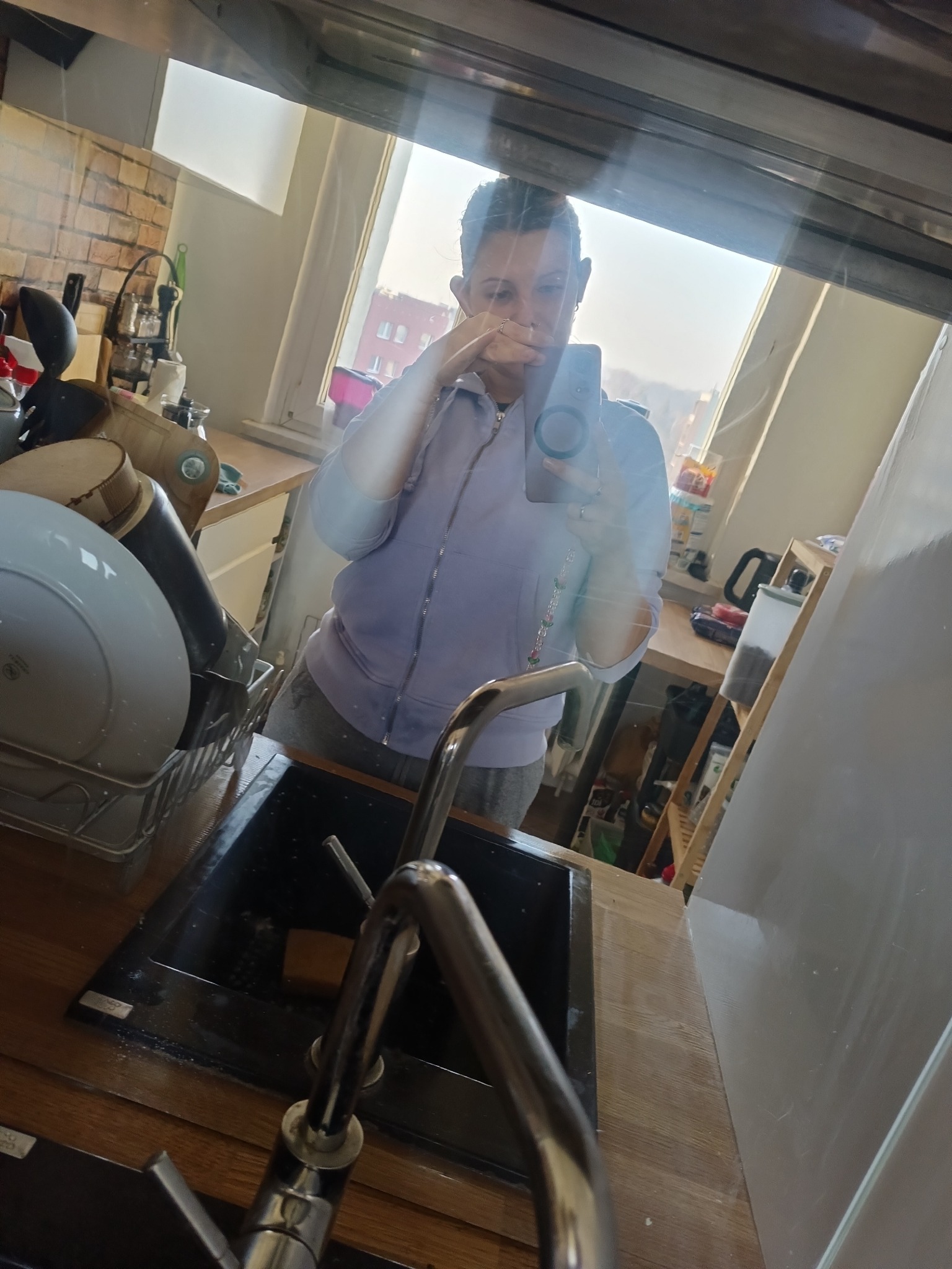 Odbicie w lustrze nad zlewem ukazuje osobę robiącą zdjęcie telefonem w kuchni z ceglaną ścianą i oknem w tle. Widoczne naczynia i akcesoria kuchenne.