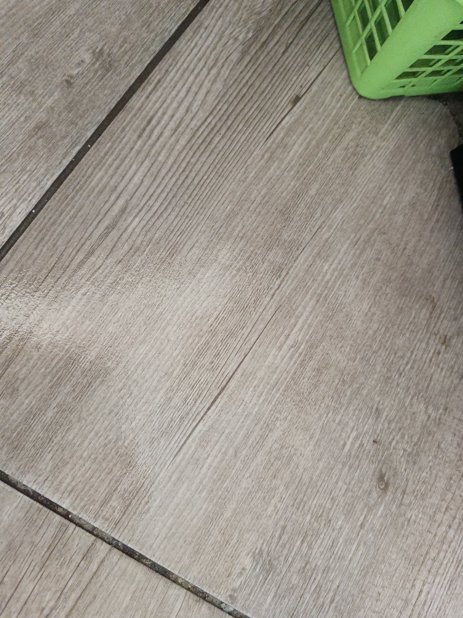 Jasne panele podłogowe imitujące drewno, ułożone ukośnie, z fragmentem zielonego koszyka w prawym górnym rogu. Widoczne fugi między panelami.