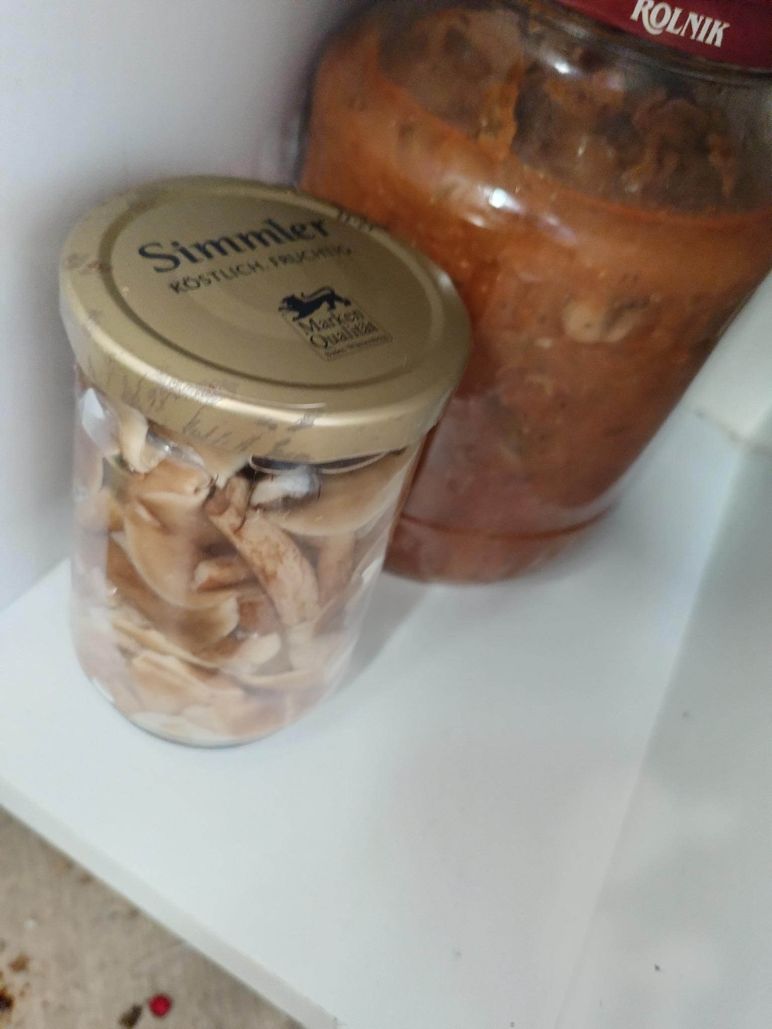 Dwa słoiki na białej półce: jeden z grzybami Simmler, drugi z sosem Rolnik. Słoik z grzybami jest w całości widoczny, a słoik z sosem jest częściowo ucięty.