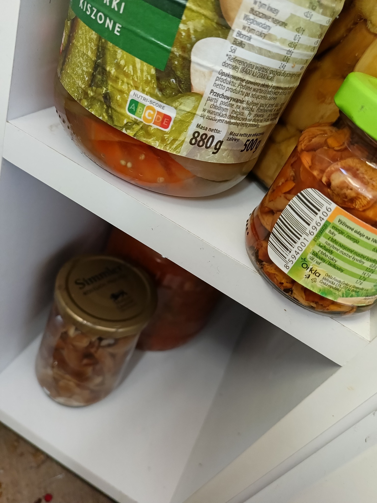 Słoiki z marynowanymi grzybami i warzywami na białej półce, widoczne etykiety z informacjami o wartościach odżywczych i składzie, ujęcie z góry pod kątem.