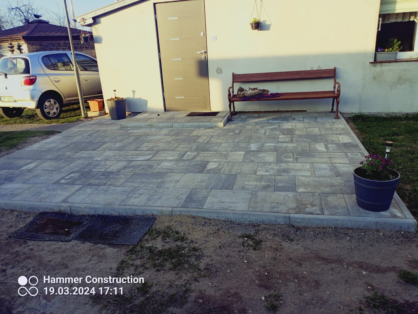 Szara kostka brukowa ułożona na posesji, obok domu z ławeczką i kotami, widok z poziomu gruntu.