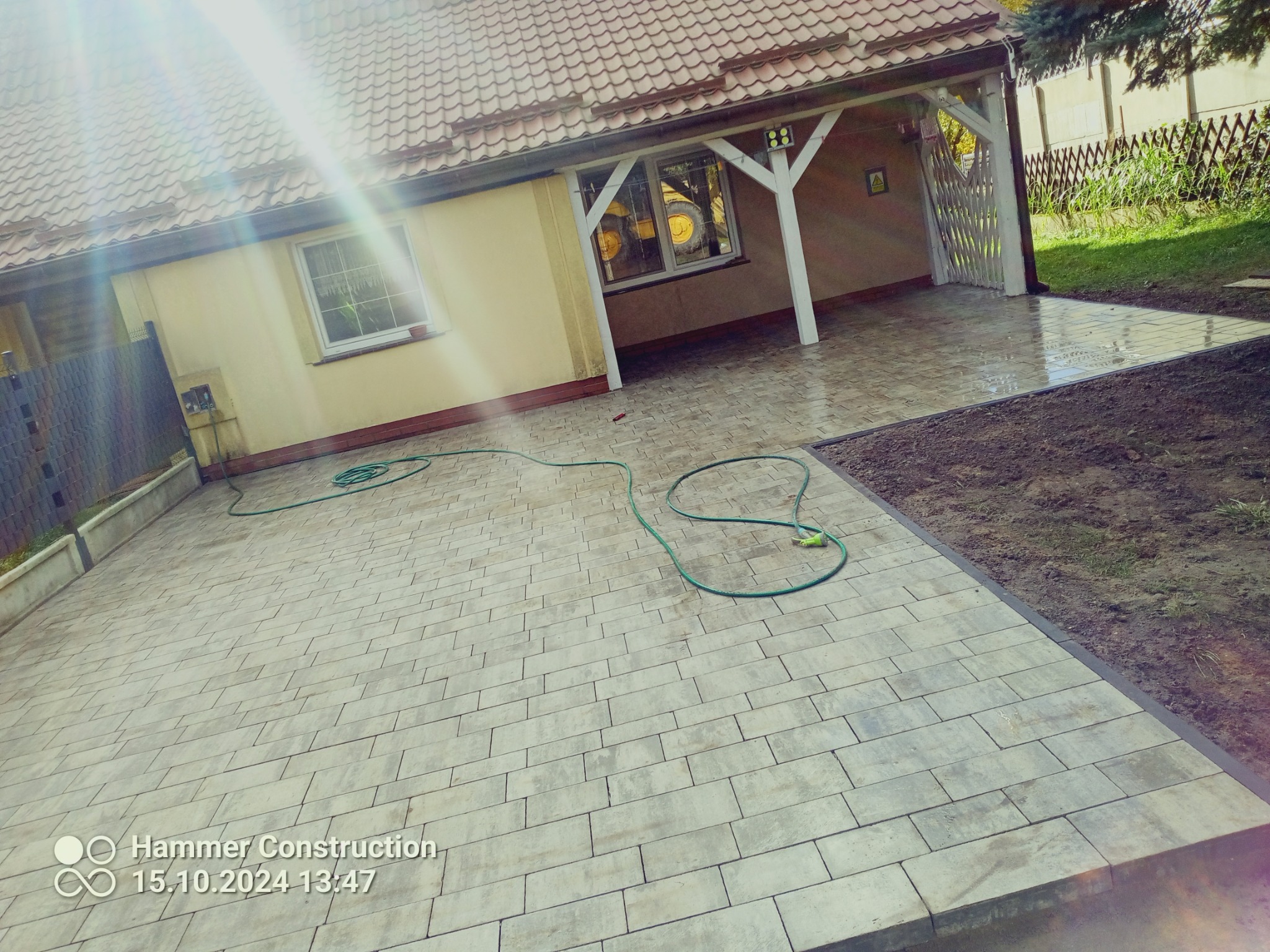Ułożona kostka brukowa przed domem z podjazdem, widoczny fragment zielonego węża ogrodowego i traktor w garażu.