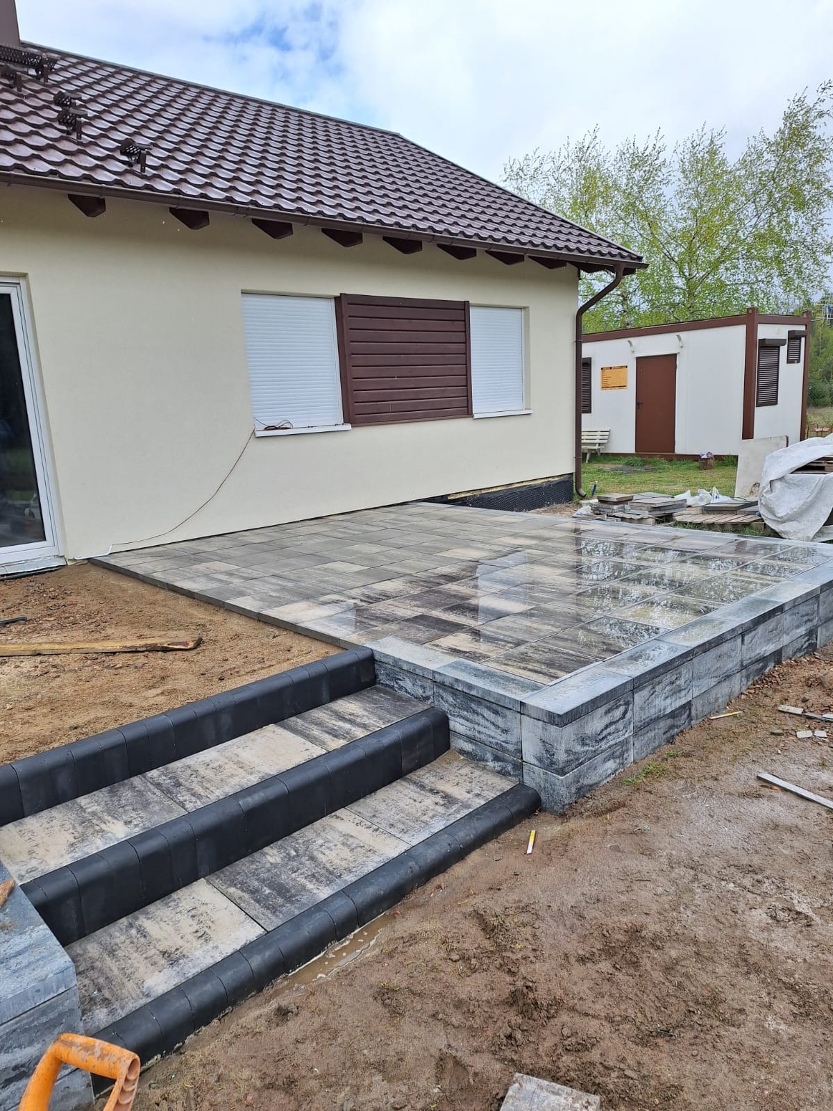 Nowo ułożony taras z kostki brukowej w odcieniach szarości, z trzema stopniami wykończonymi czarnym obrzeżem, przylegający do jasnego domu z brązowym dachem i roletami.