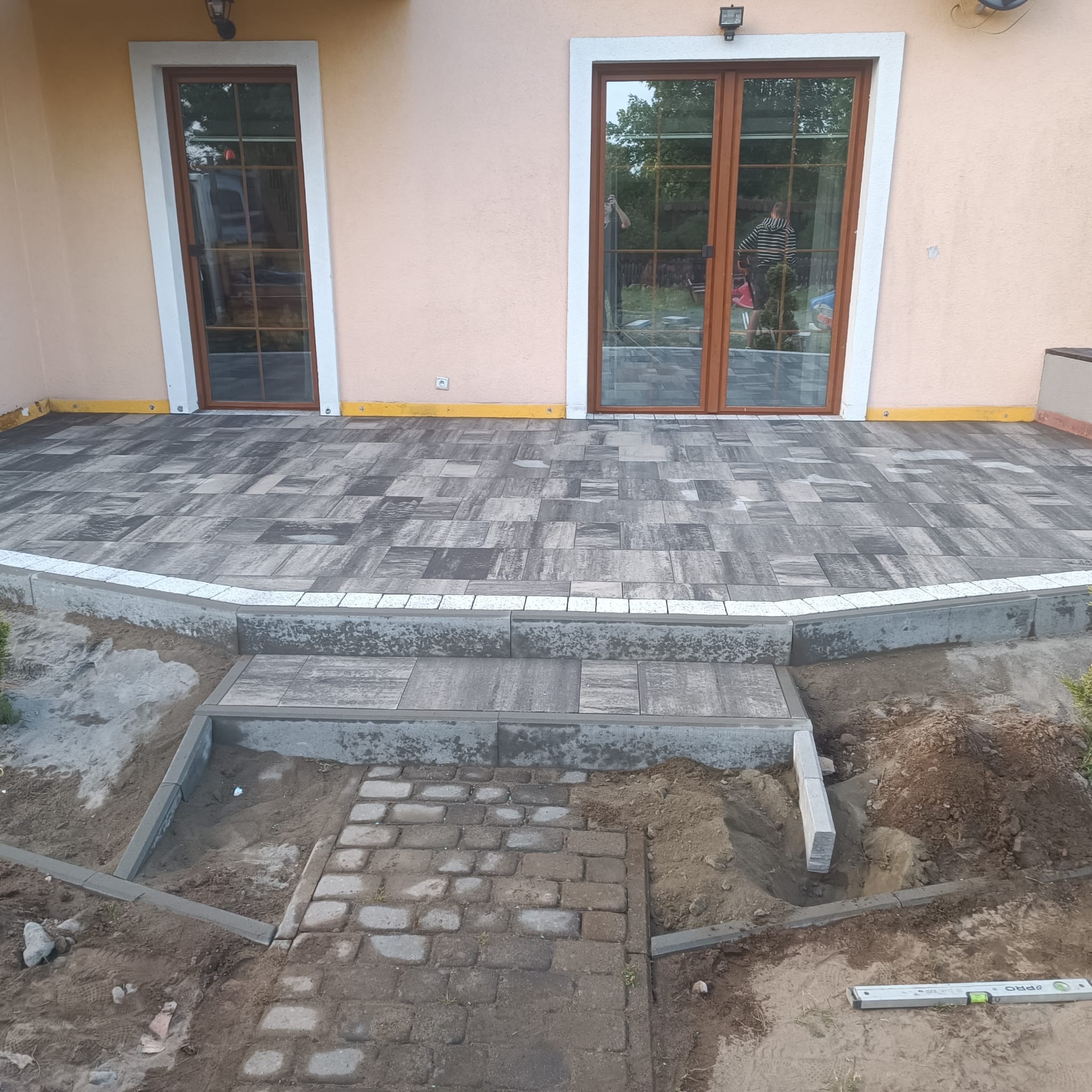 Nowo ułożony taras z kostki brukowej w odcieniach szarości, z widocznymi schodami i obrzeżami, częściowo w trakcie prac ziemnych i przygotowania terenu.