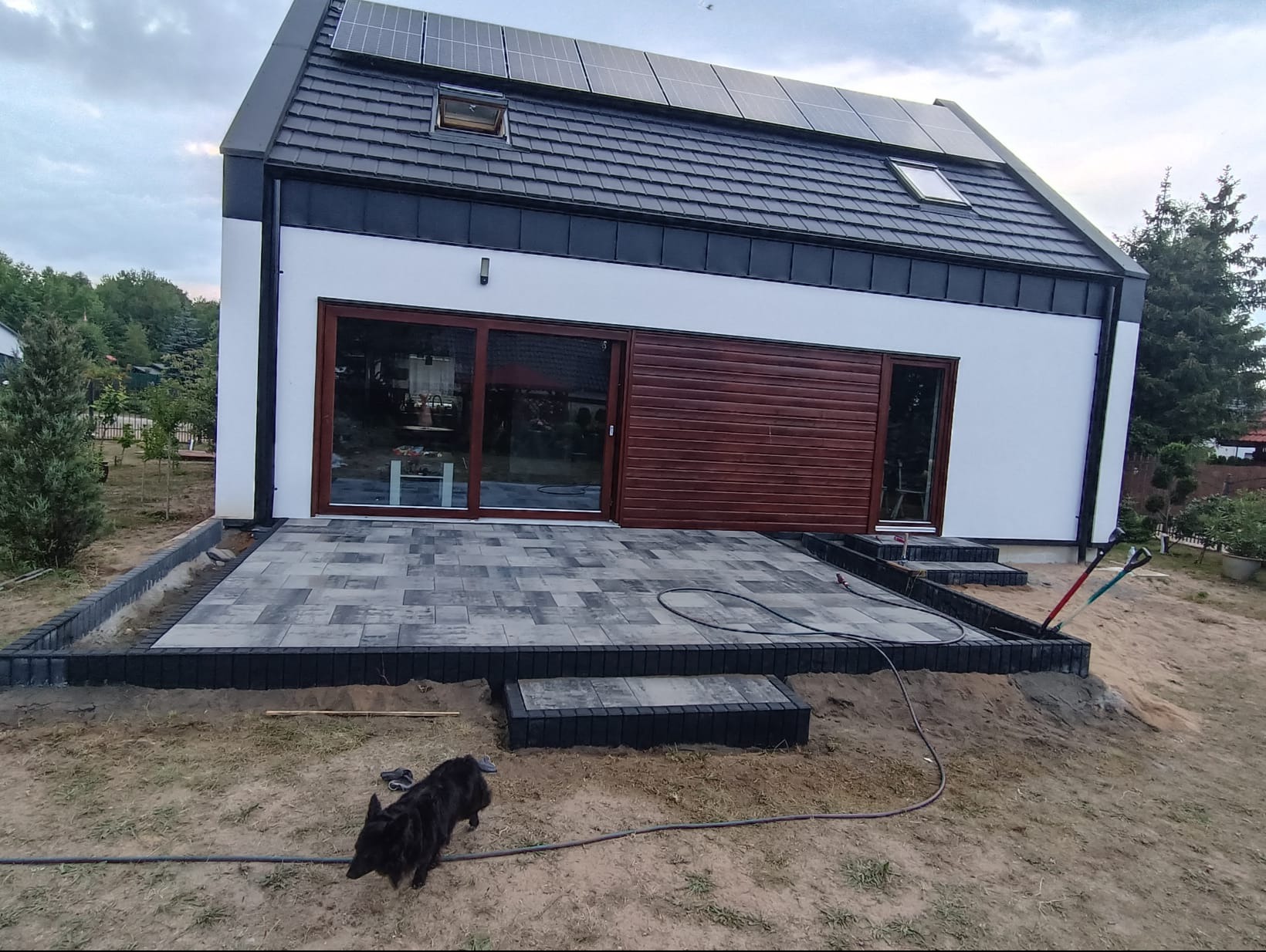 Nowoczesny dom z szarym brukowanym tarasem i czarną obwódką; czarny pies na pierwszym planie, narzędzia ogrodnicze oparte o krawędź tarasu.