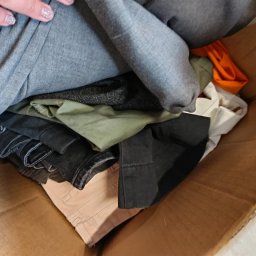 Sole Agata Zielińska - Kartonowe pudło wypełnione różnorodnymi ubraniami, w tym szare bluzy, zielone spodnie i czarne jeansy, z widoczną dłonią z brokatowym manicure.