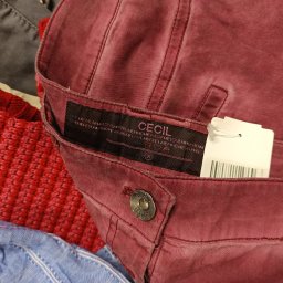 Sole Agata Zielińska - Burgundowe spodnie marki Cecil z metką, leżące na stercie innych ubrań, widoczne fragmenty czerwonego swetra i niebieskiej koszuli.