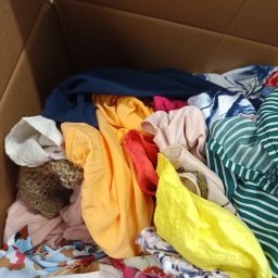 Sole Agata Zielińska - Pudełko kartonowe wypełnione różnorodną, kolorową odzieżą damską, w tym bluzki w paski, wzorzyste tkaniny i dzianinowy sweter, z widoczną dłonią w dolnym rogu.