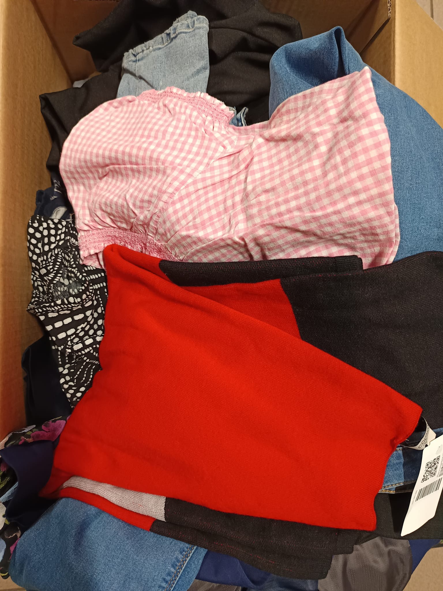 Karton wypełniony różnorodną odzieżą używaną, w tym bluzka w różową kratkę, czerwony sweter i dżinsowe ubrania.