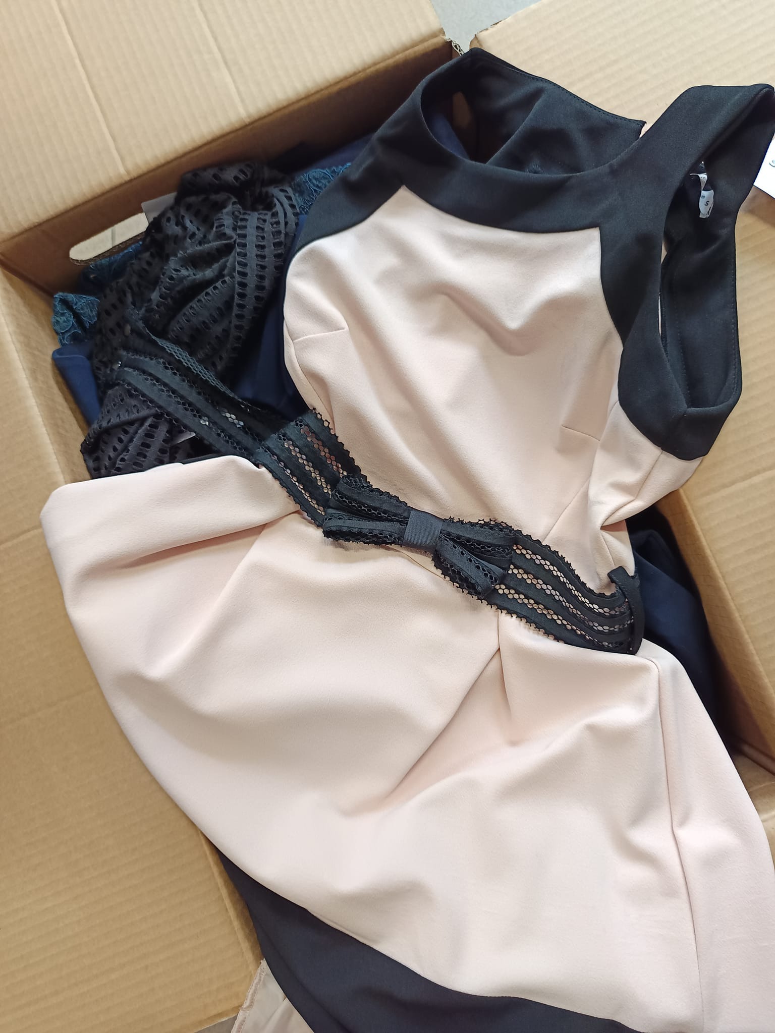 Otwarty karton wypełniony odzieżą damską, w tym sukienka w kolorze pudrowego różu z czarnymi detalami i koronkową aplikacją, oraz inne ubrania w odcieniach granatu i czerni.