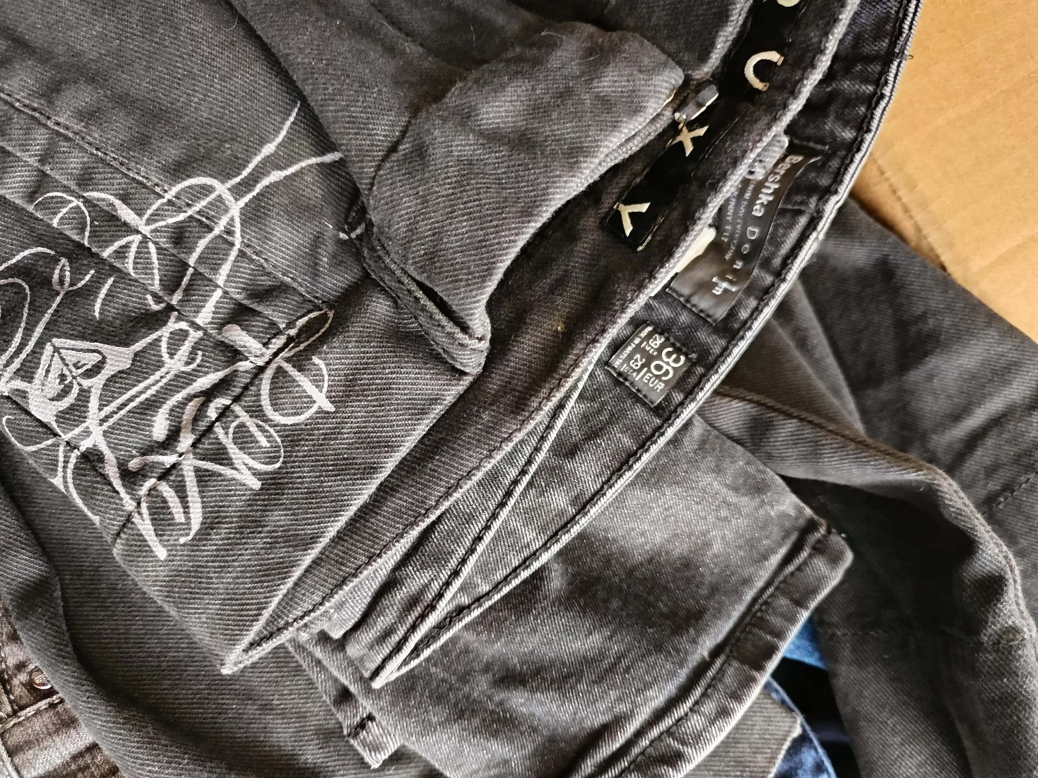 Poskładane czarne jeansy z białym, abstrakcyjnym nadrukiem i widoczną metką marki Bershka Denim.