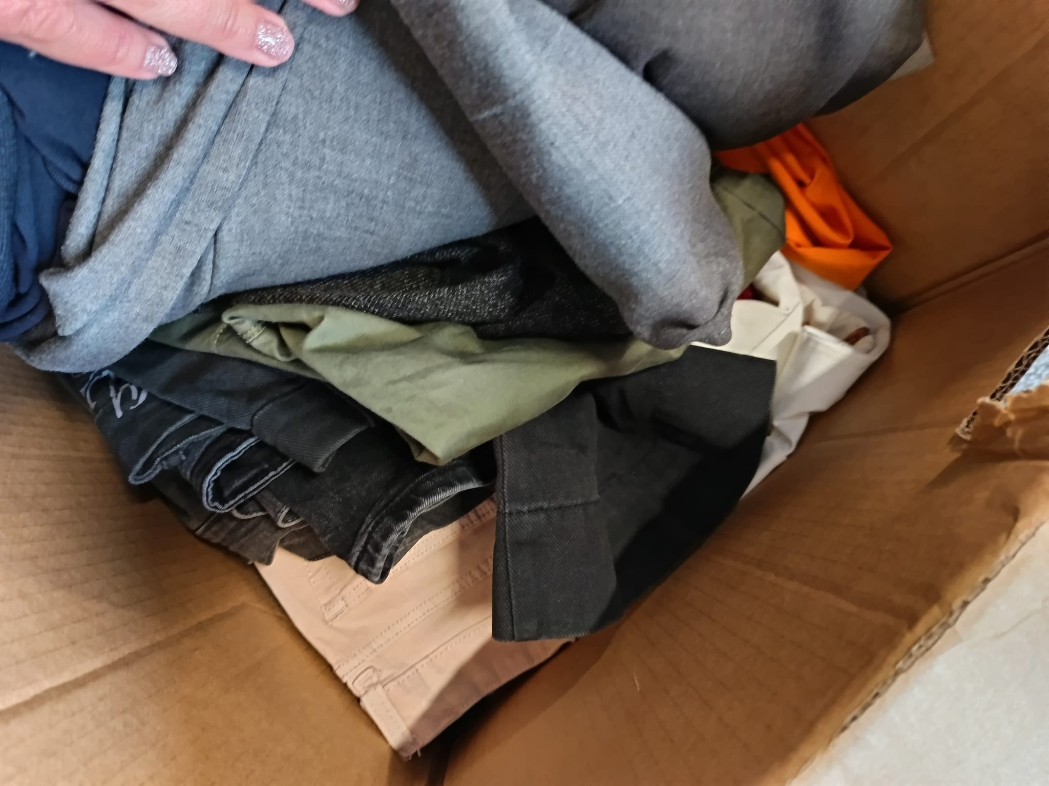 Kartonowe pudło wypełnione różnorodnymi ubraniami, w tym szare bluzy, zielone spodnie i czarne jeansy, z widoczną dłonią z brokatowym manicure.