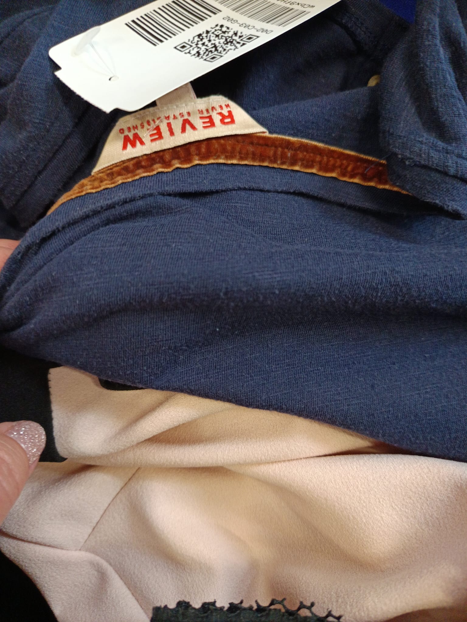 Niebieska koszulka z brązowym welurowym wykończeniem dekoltu i metką marki Review, leżąca na jasnobeżowej bluzce.