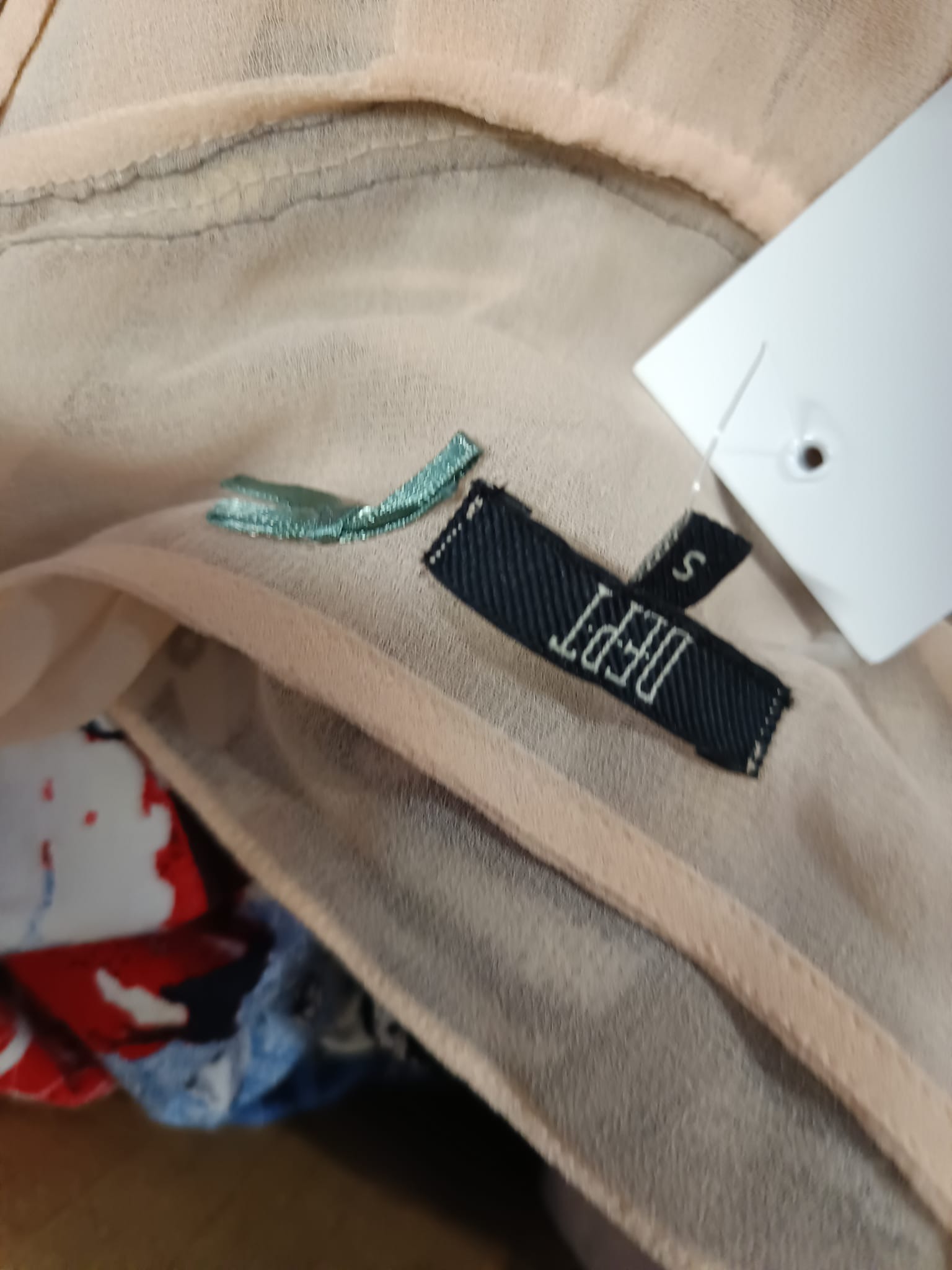 Bluzka z delikatnego, beżowego materiału z metką marki DEPT w rozmiarze S i fragmentem haftu w kolorze turkusowym, widoczna część kolorowej odzieży w tle.