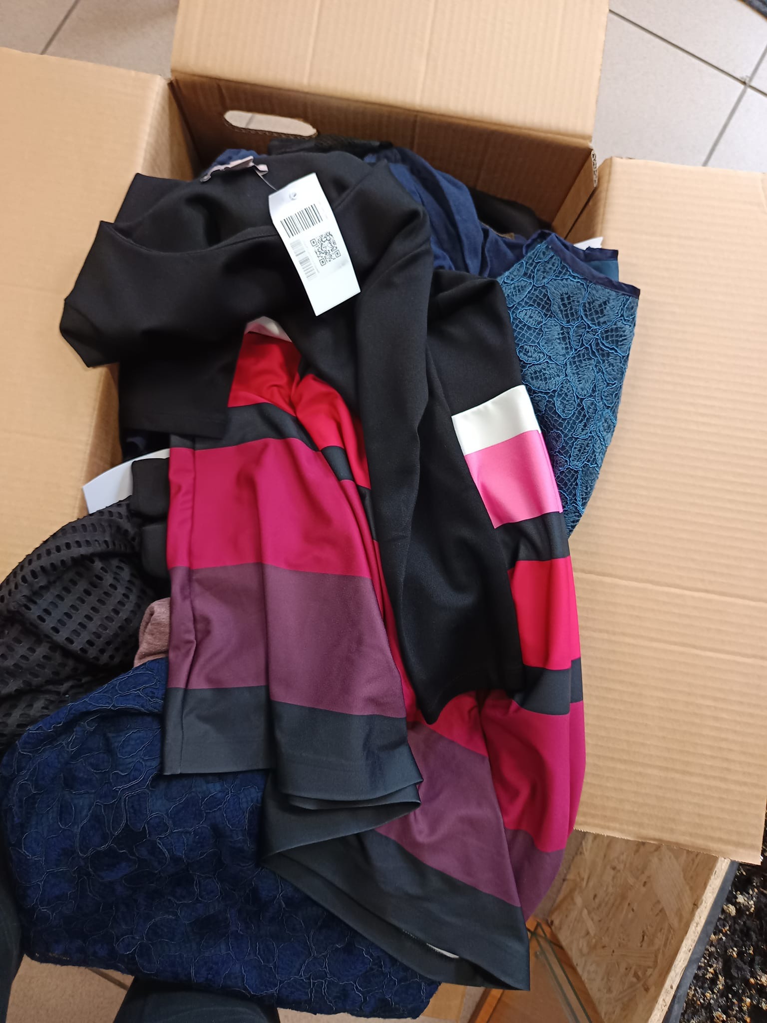 Kartonowe pudło wypełnione różnorodną odzieżą damską, w tym czarną sukienką z metką, bluzką w kolorowe pasy i granatową koronkową spódnicą.