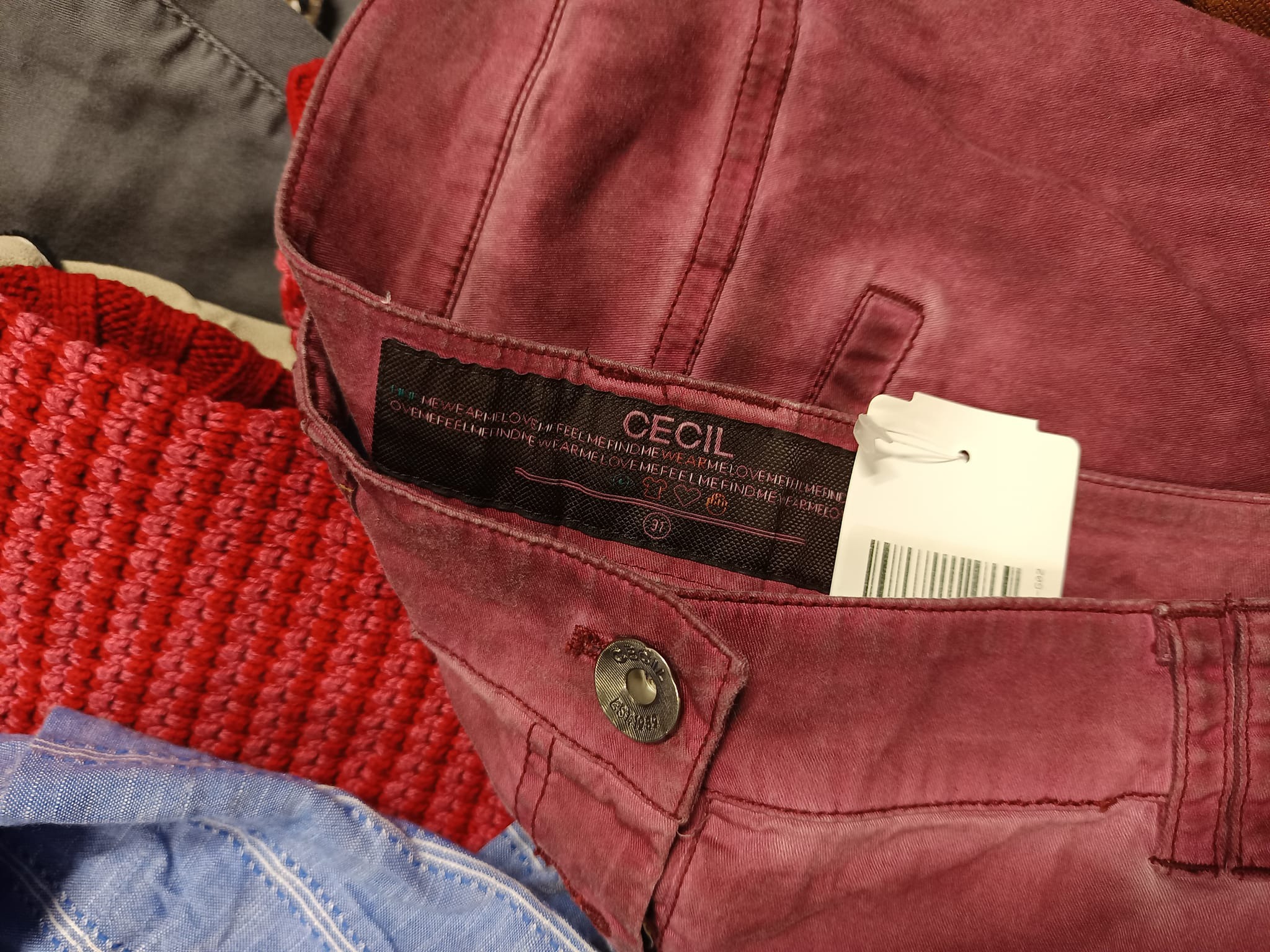 Burgundowe spodnie marki Cecil z metką, leżące na stercie innych ubrań, widoczne fragmenty czerwonego swetra i niebieskiej koszuli.