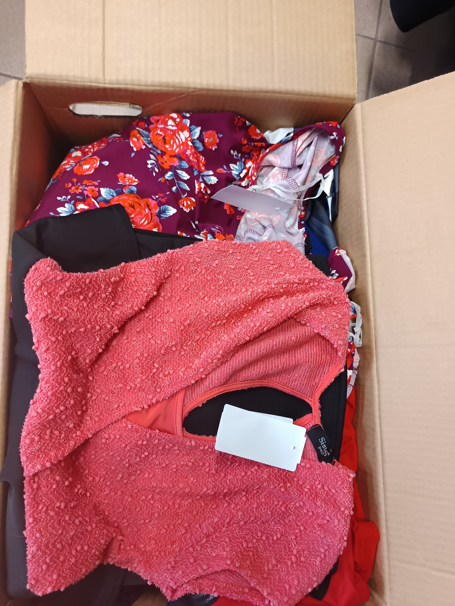 Otwarty karton wypełniony damską odzieżą w różnych kolorach i wzorach, widoczna bluzka w kolorze koralowym z metką oraz sukienka w kwiaty.