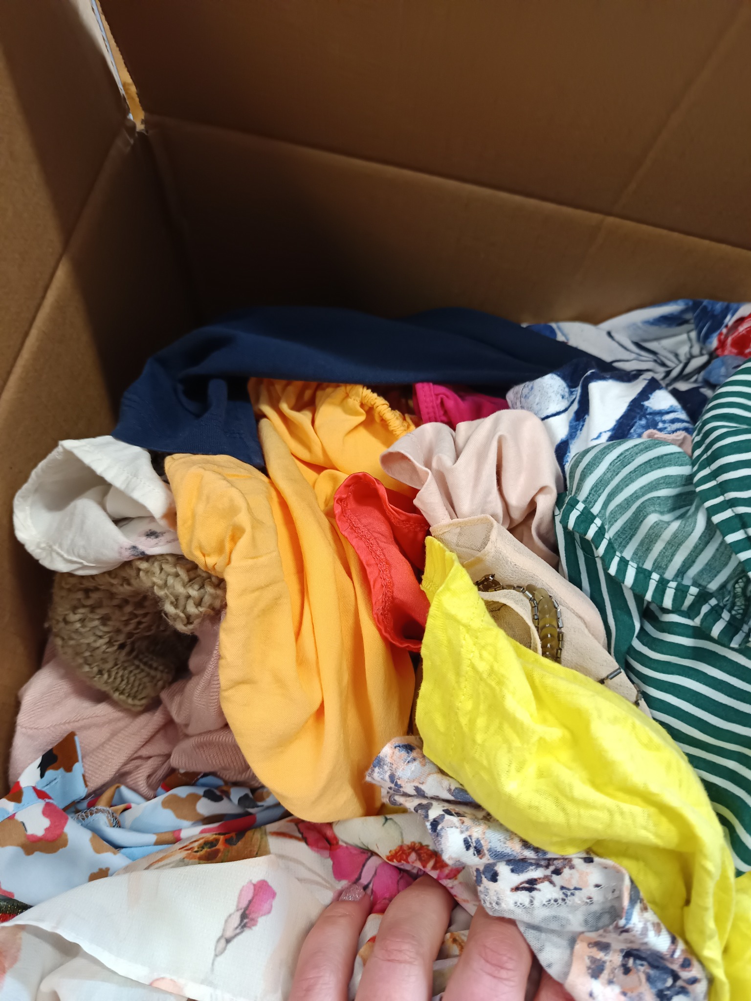 Pudełko kartonowe wypełnione różnorodną, kolorową odzieżą damską, w tym bluzki w paski, wzorzyste tkaniny i dzianinowy sweter, z widoczną dłonią w dolnym rogu.