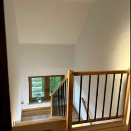 Nowoczesne schody drewniane z czarnymi balustradami, widok z góry, na podłodze niebieska taśma zabezpieczająca, w tle okno z widokiem na zieleń.