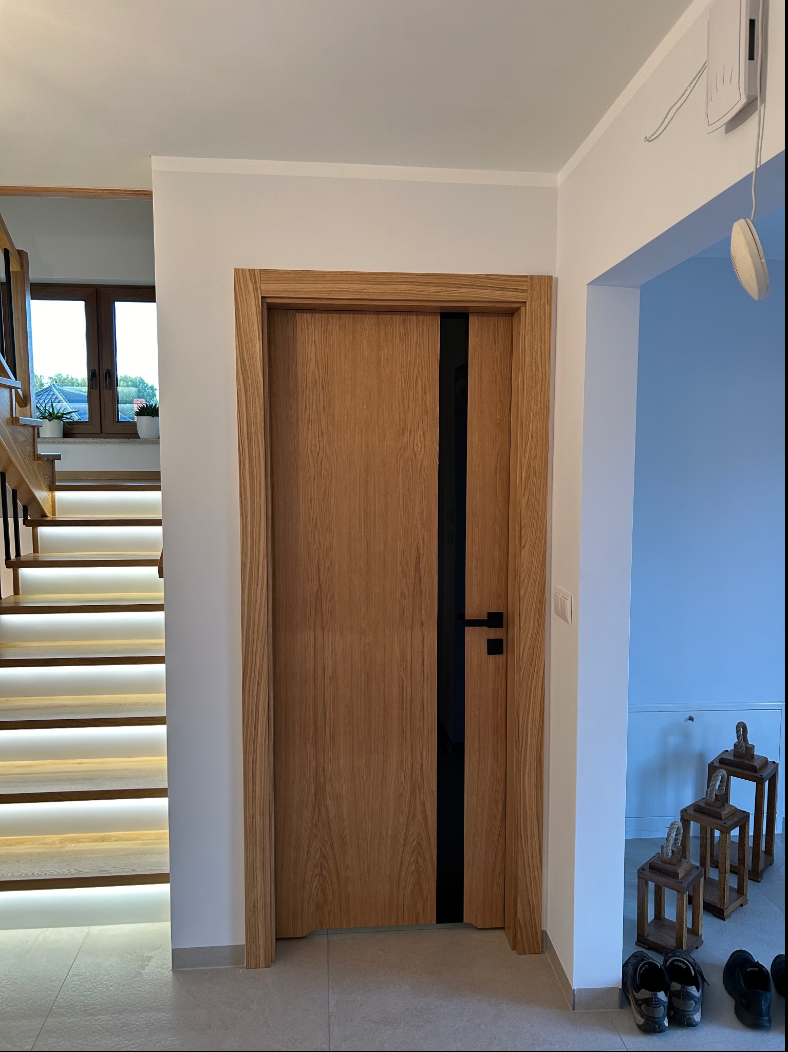 Drewniane drzwi wewnętrzne z czarnym pionowym akcentem, widoczne z perspektywy korytarza z podświetlanymi schodami i dekoracyjnymi lampionami.