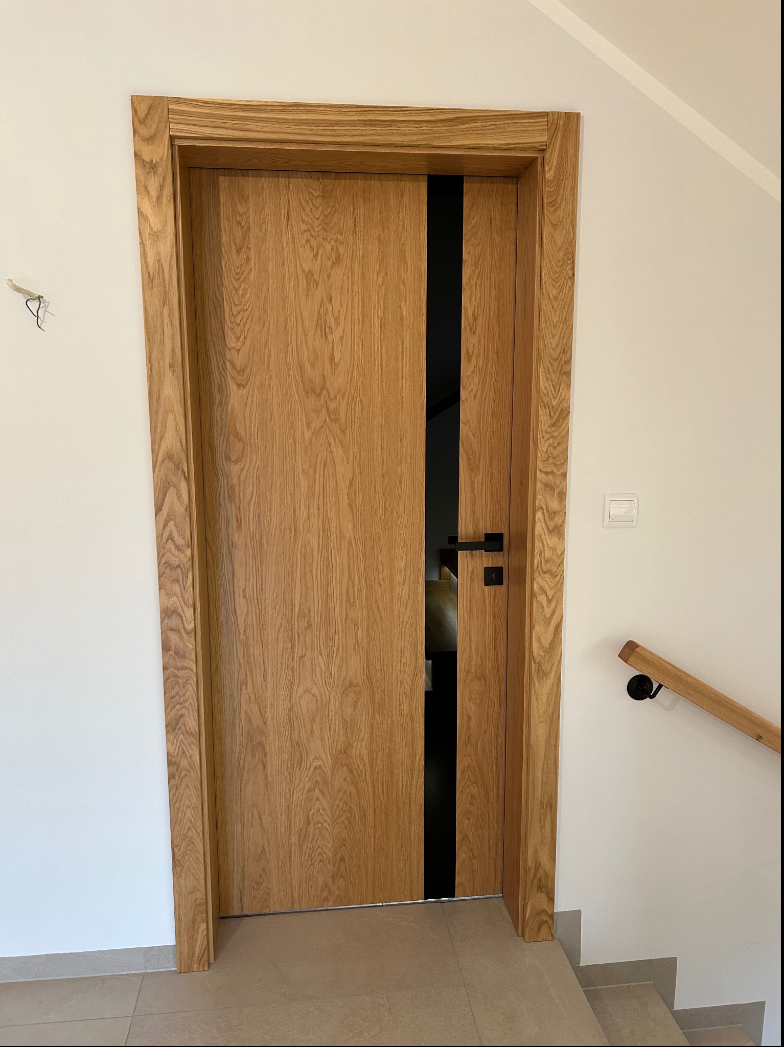 Dębowa ościeżnica z drewnianymi drzwiami z czarnym pionowym panelem, czarna klamka na tle białej ściany i schodów z drewnianą barierką.