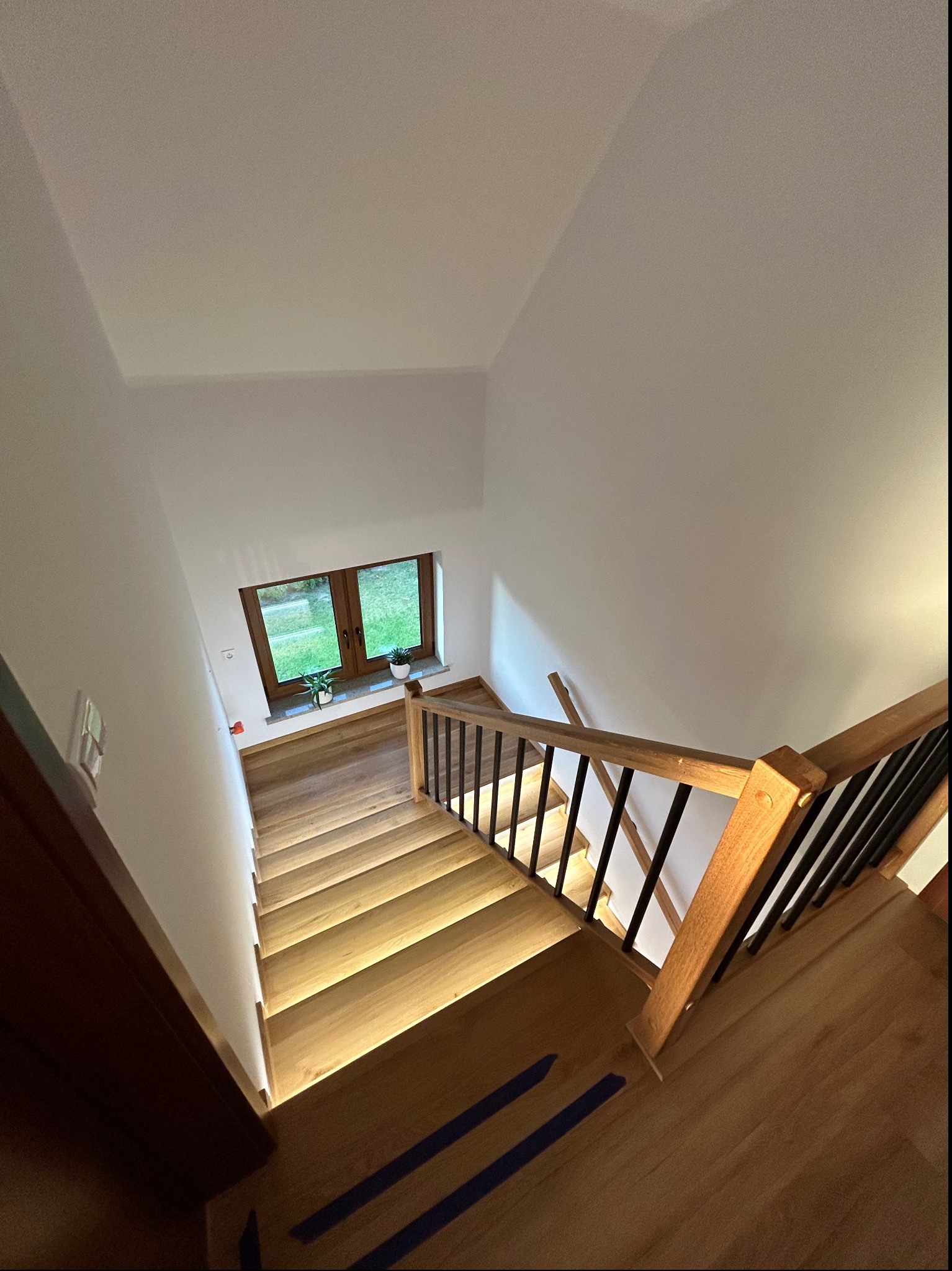 Drewniane schody z podświetleniem LED, widok z góry, czarne metalowe balustrady, okno z widokiem na zieleń w tle, minimalistyczne wnętrze.