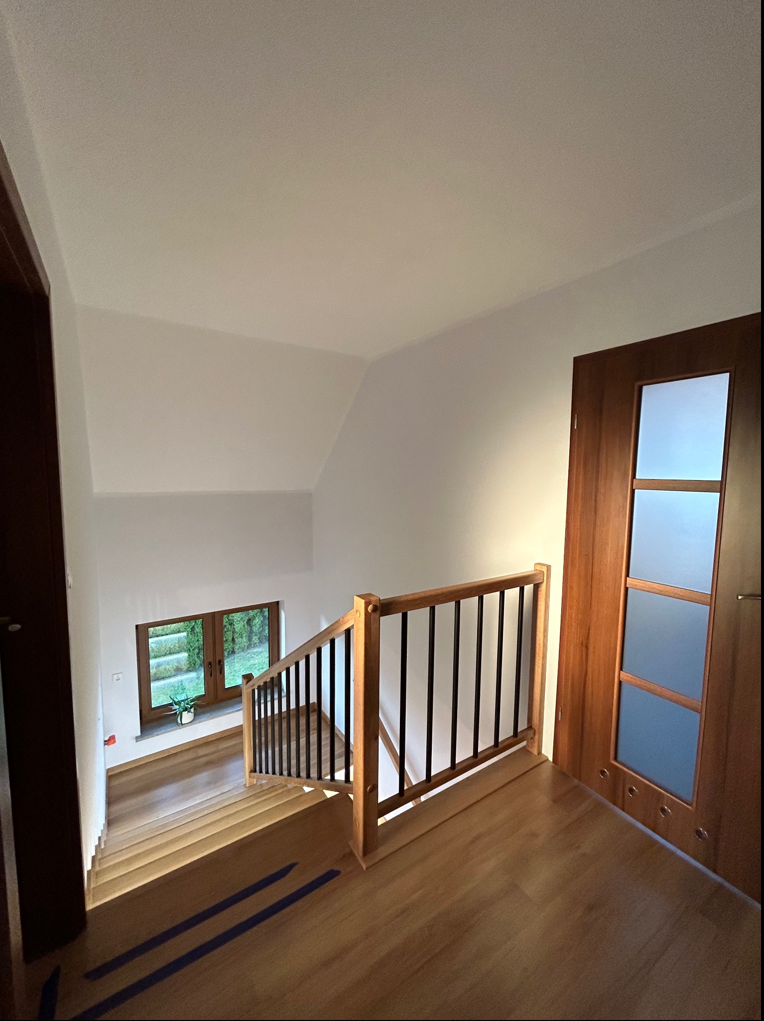 Wnętrze domu z drewnianymi schodami z czarnymi balustradami, widok z góry na dół, z oknem w tle i drewnianymi drzwiami po prawej stronie.