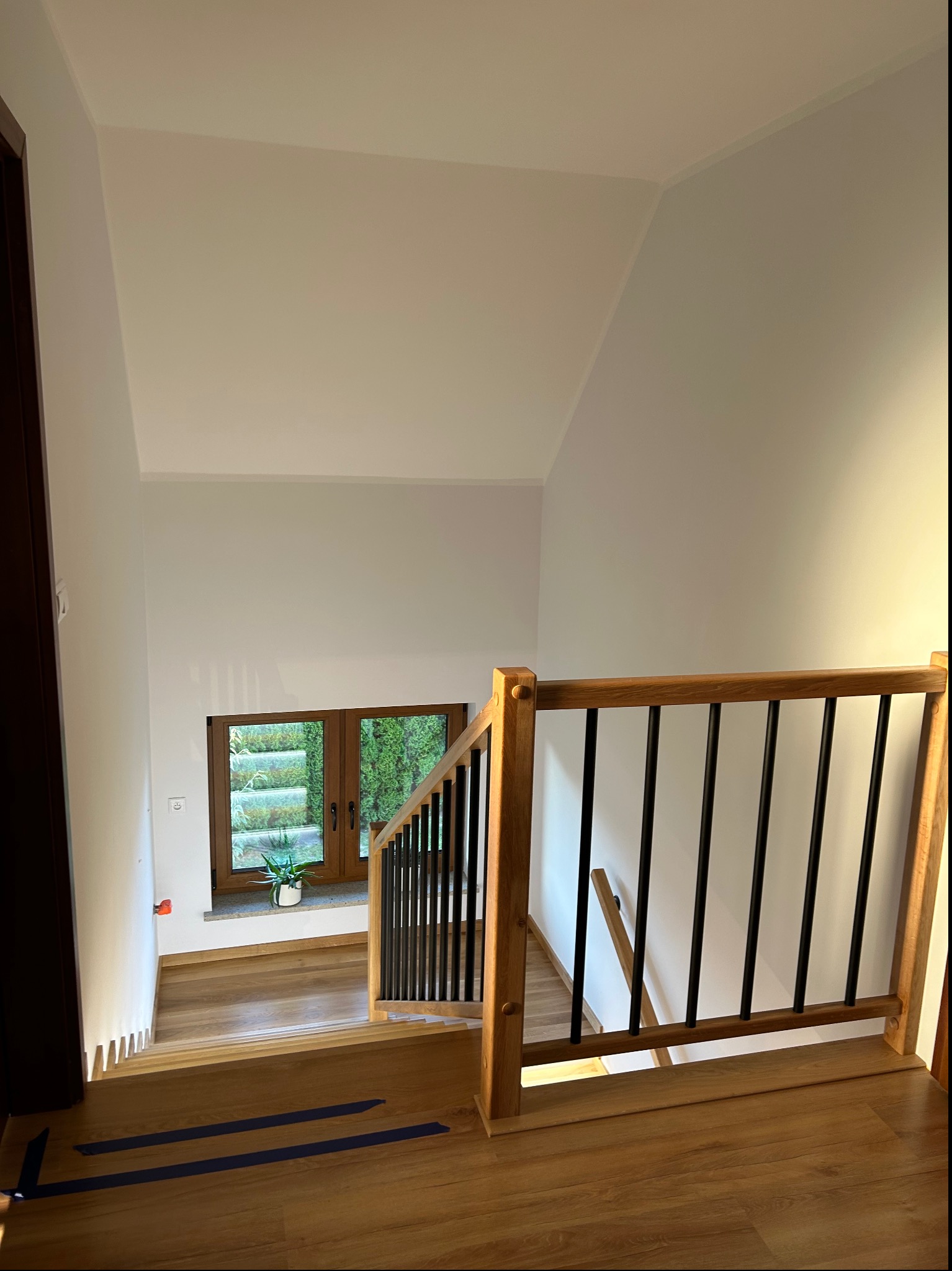Nowoczesne schody drewniane z czarnymi balustradami, widok z góry, na podłodze niebieska taśma zabezpieczająca, w tle okno z widokiem na zieleń.