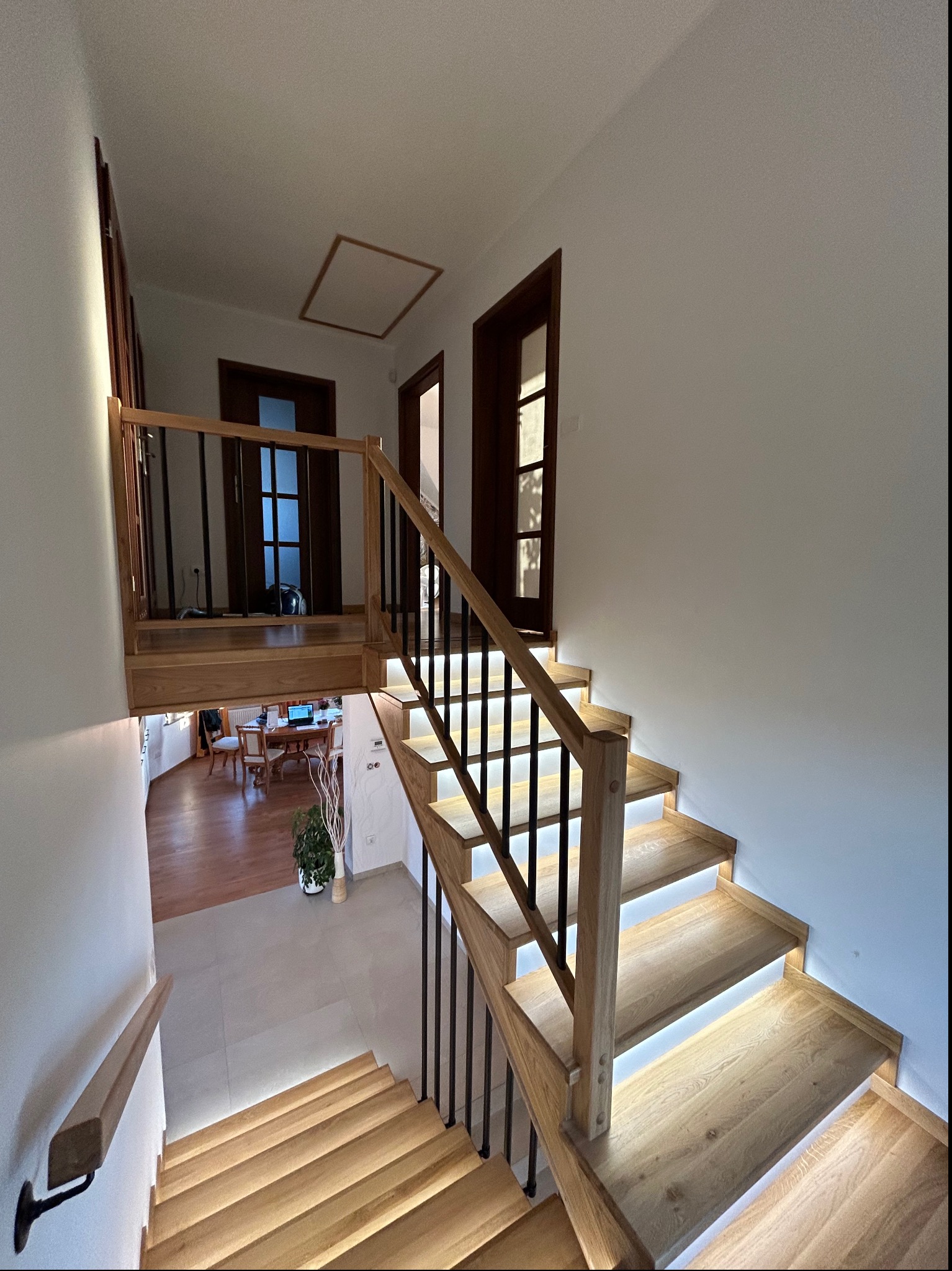 Drewniane schody z podświetleniem LED, czarne metalowe balustrady, widok z góry na otwartą przestrzeń z drewnianą podłogą i stołem. W tle widoczne drzwi i mała roślina w doniczce.