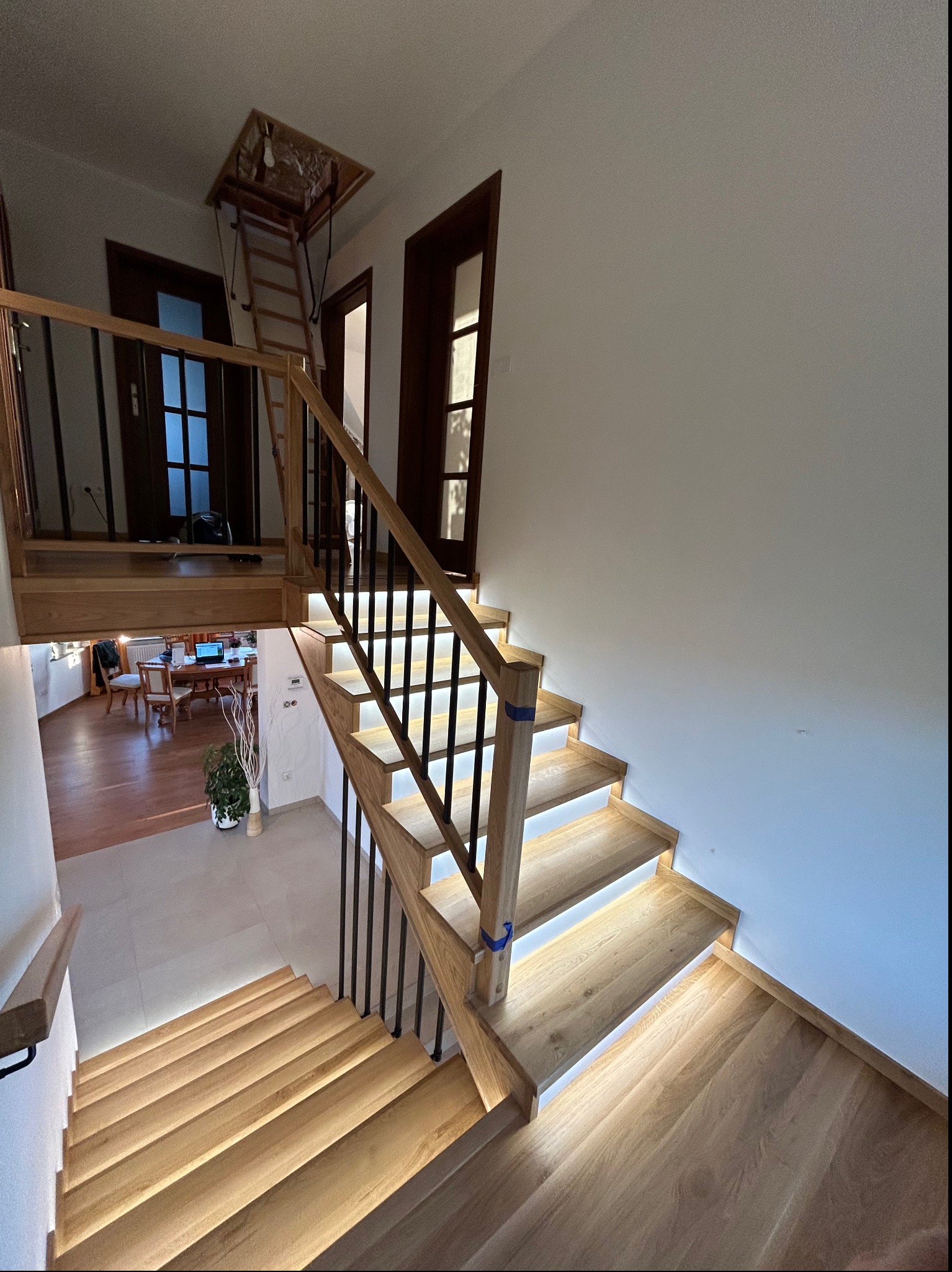 Drewniane schody z podświetleniem LED, czarne metalowe balustrady, widok na antresolę z drabinką i otwarte drzwi do innych pomieszczeń.