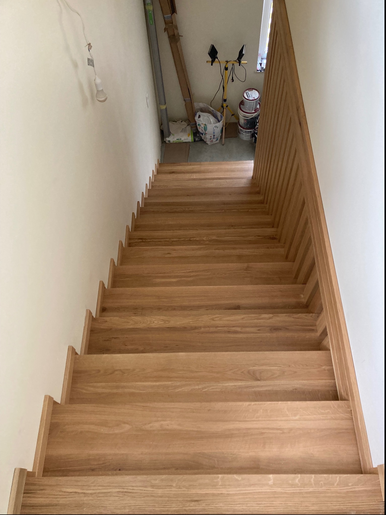 Widok z góry na drewniane schody o jasnym odcieniu, z minimalistyczną balustradą po prawej stronie, w tle widoczne narzędzia i materiały budowlane.