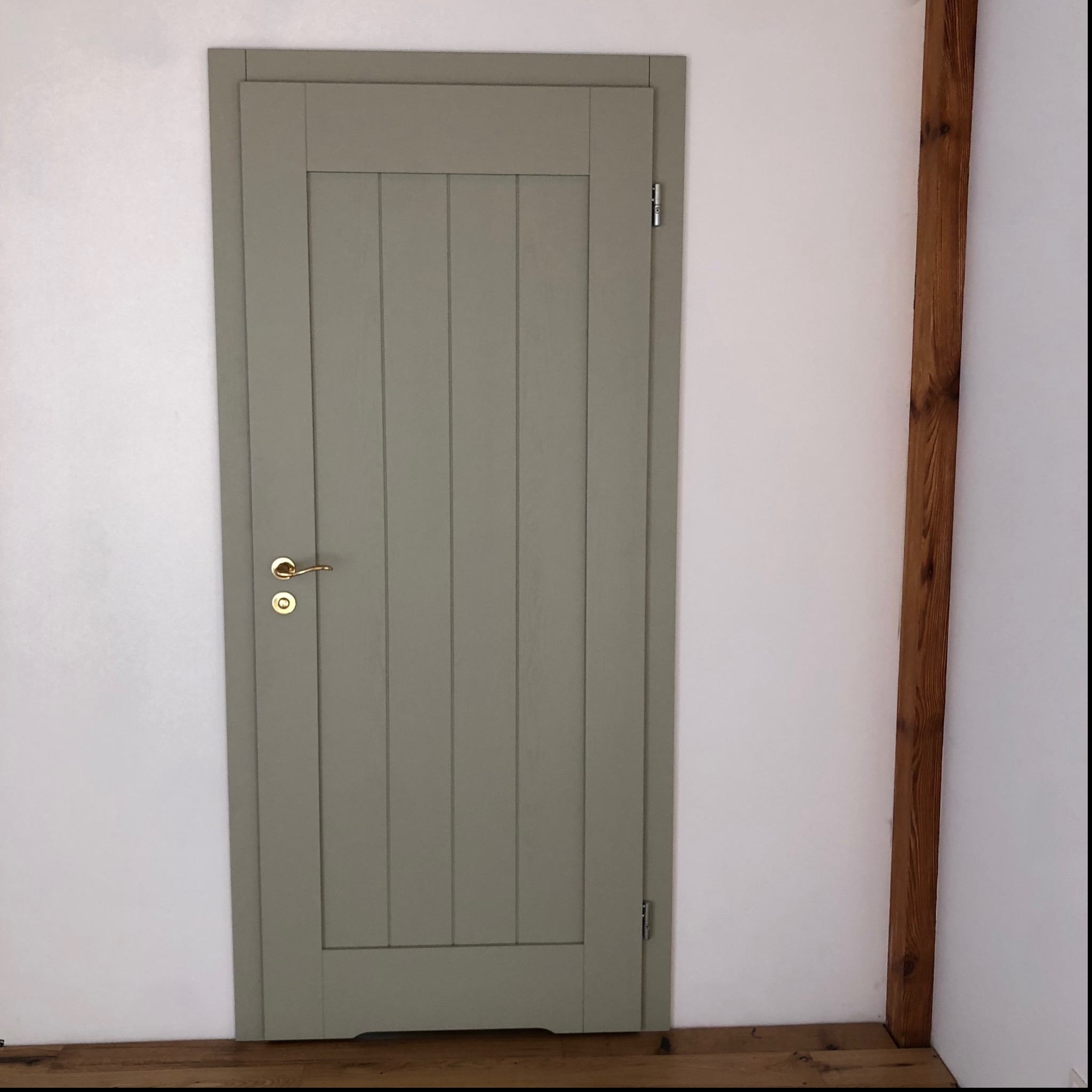 Szara, drewniana, wewnętrzna drzwi z pionowymi panelami, złotą klamką i widoczną drewnianą ramą w rogu pomieszczenia na białym tle.
