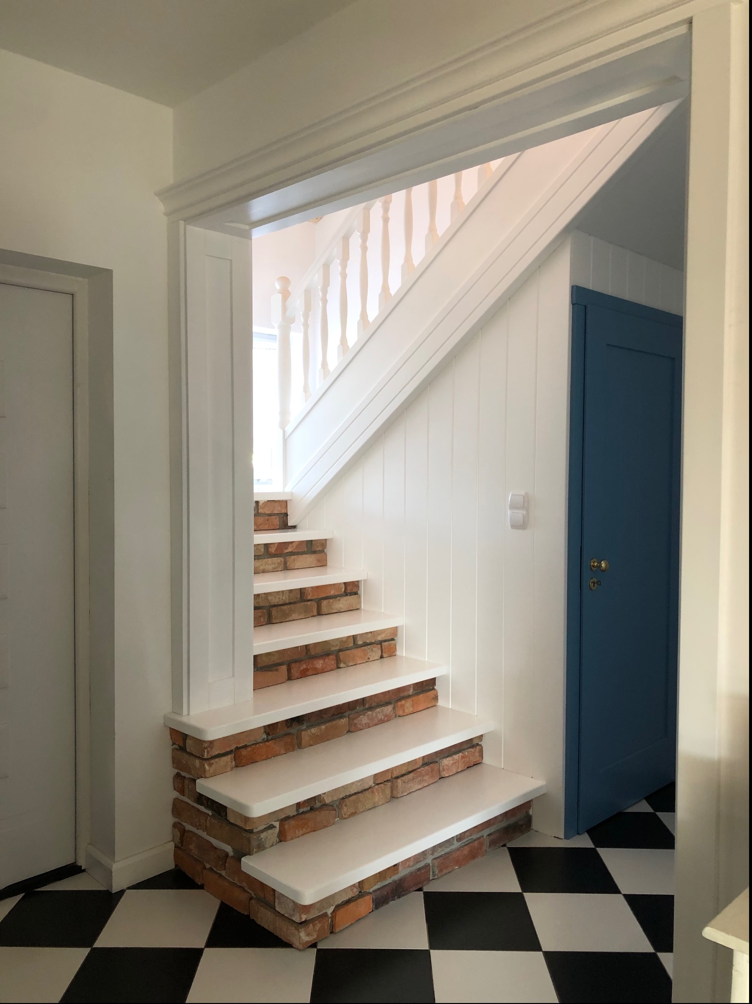 Białe schody z ceglanymi wstawkami w nowoczesnym wnętrzu z szachownicą na podłodze i niebieskimi drzwiami.