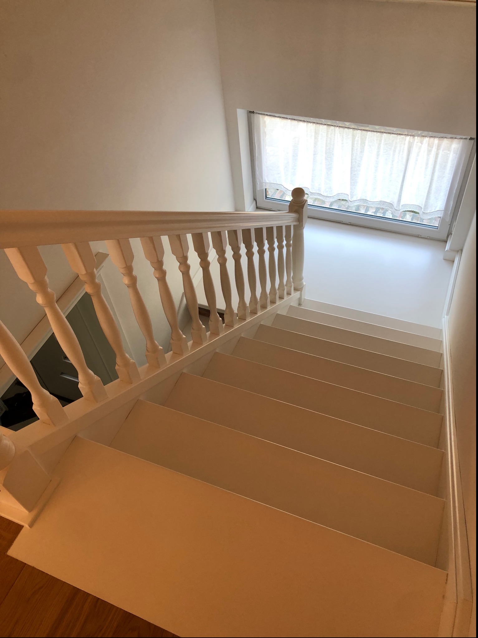 Białe, drewniane schody prowadzące w dół, oświetlone światłem z okna z koronkową firanką, widoczne elementy drewnianej konstrukcji schodów i fragment podłogi na dole.