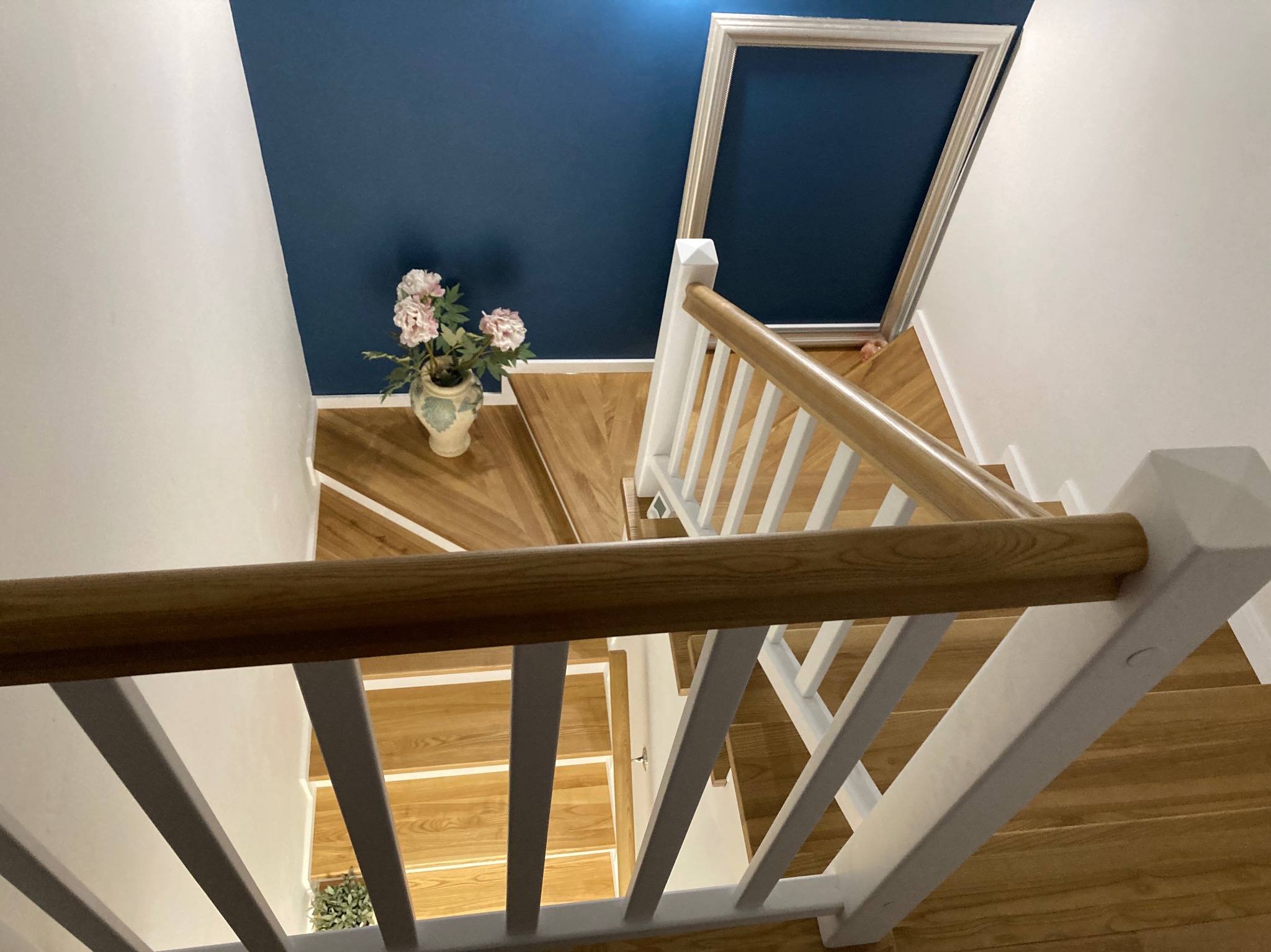 Widok z góry na drewniane schody z białymi balustradami, na podeście wazon z kwiatami, ściana w kolorze granatowym z ramą.