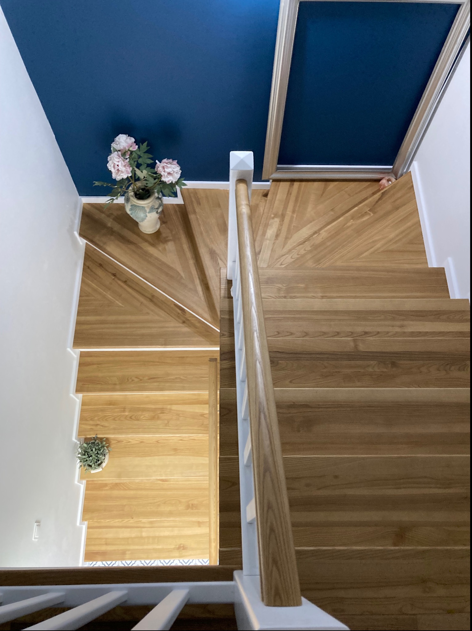 Schody drewniane z jasnego drewna, widok z góry, z białymi barierkami i niebieskim sufitem, dekorowane kwiatami w wazonie i małą rośliną doniczkową.