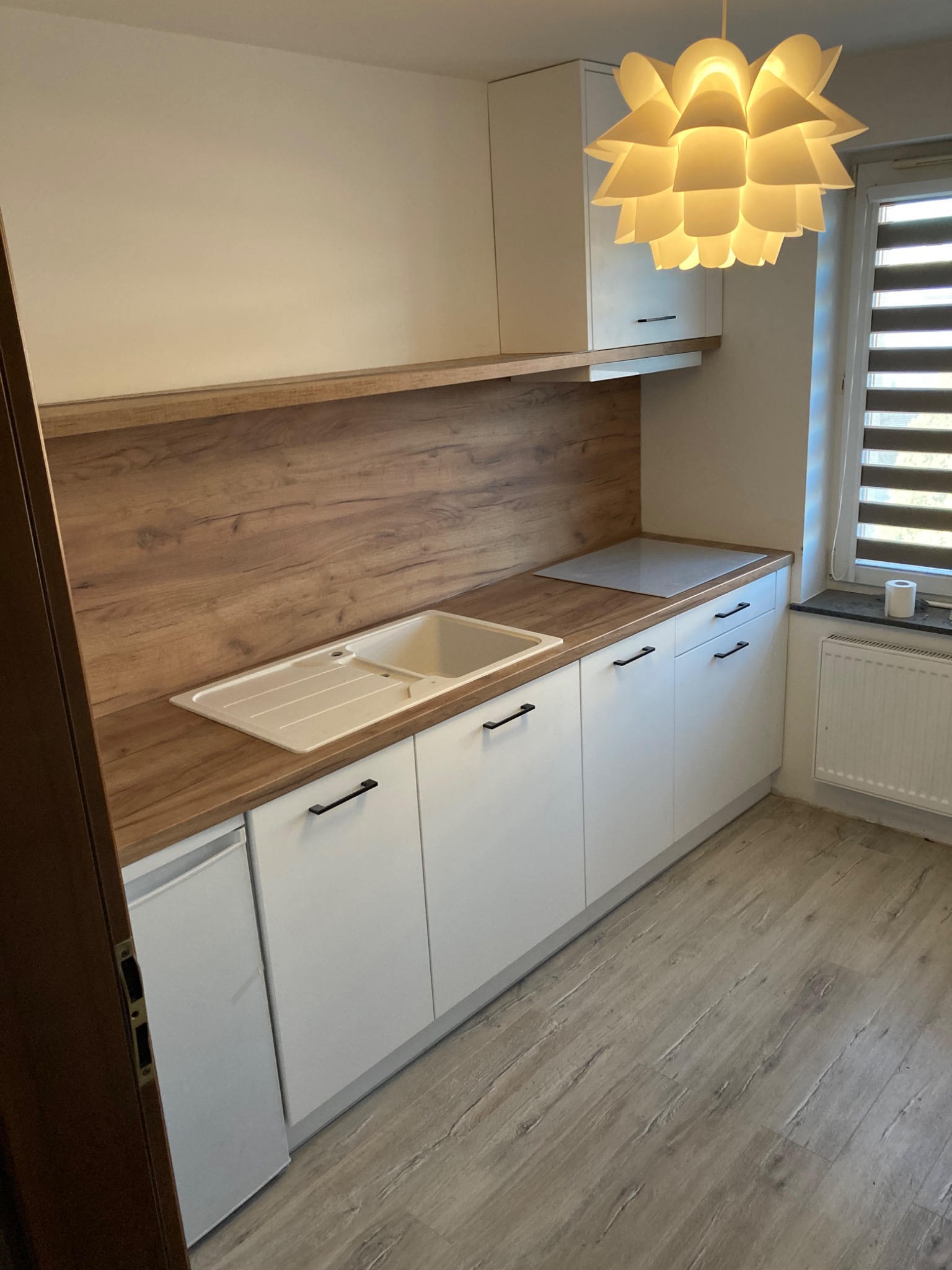 Nowoczesna kuchnia z białymi szafkami, drewnianym blatem i panelem ściennym, oświetlona designerską lampą, z widocznym fragmentem okna z żaluzjami.