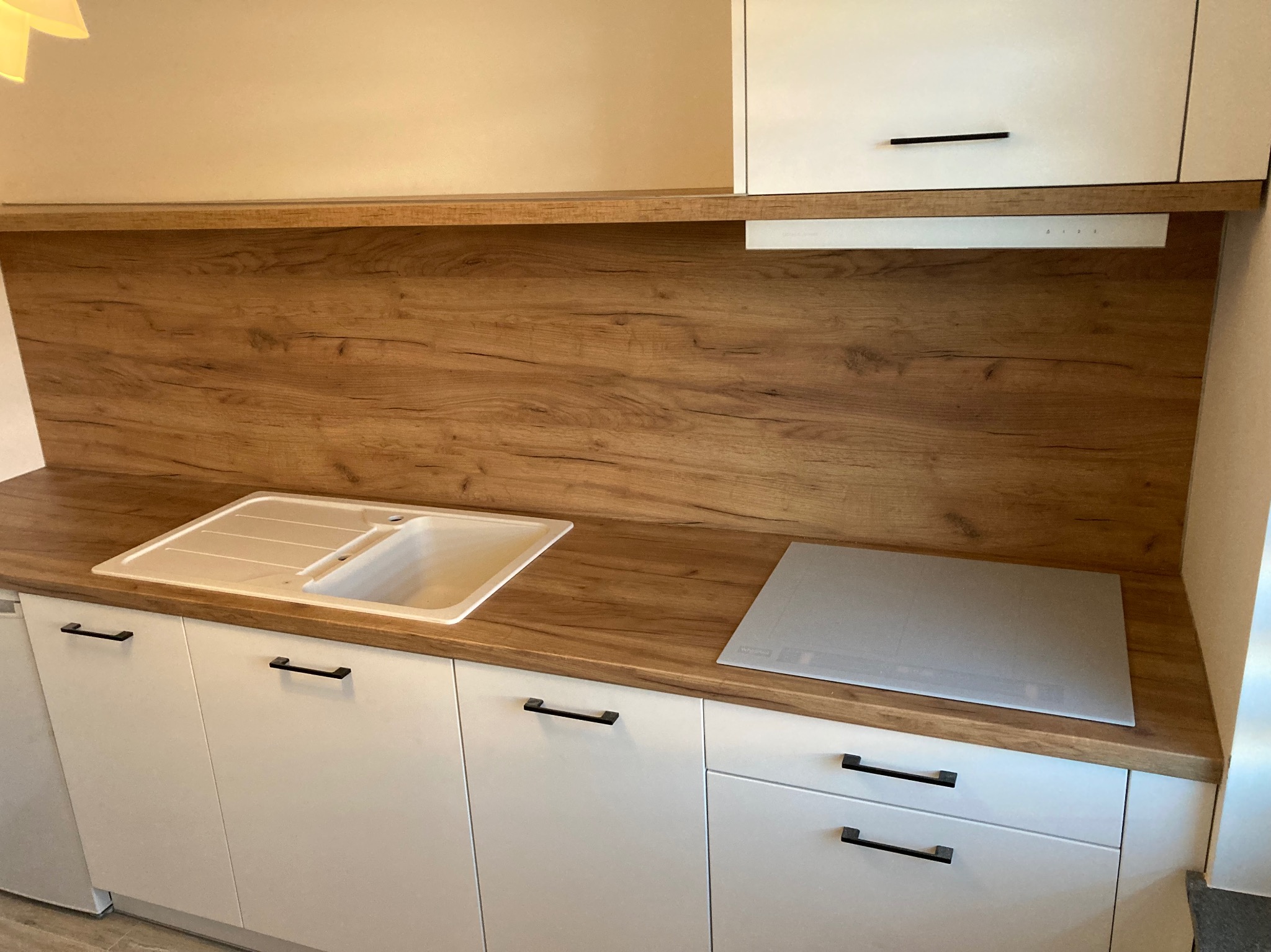 Fragment zabudowy kuchennej: białe szafki z czarnymi uchwytami, drewniany blat i panel nad blatem, zlewozmywak z ociekaczem oraz płyta indukcyjna.
