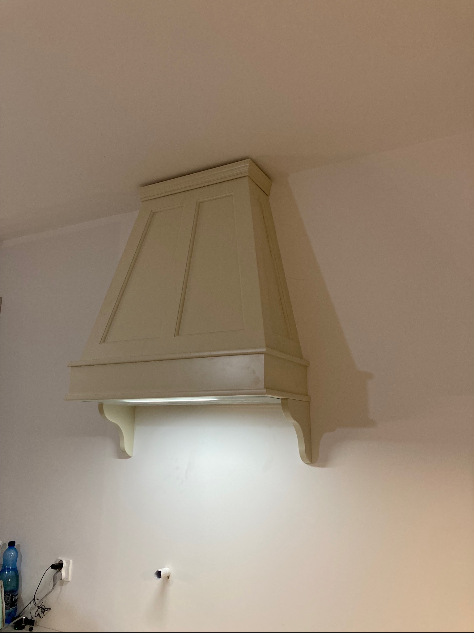 Kremowy okap kuchenny w stylu rustykalnym zamontowany na ścianie w Kamienicy Królewskiej, z widocznym oświetleniem LED pod spodem. Na ścianie widoczne gniazdka elektryczne i rura.